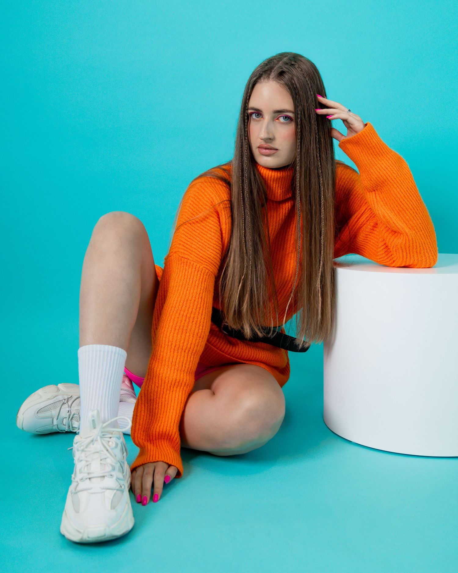 Young woman in orange turtleneck sweater dress and white sneakers posing against a teal background.