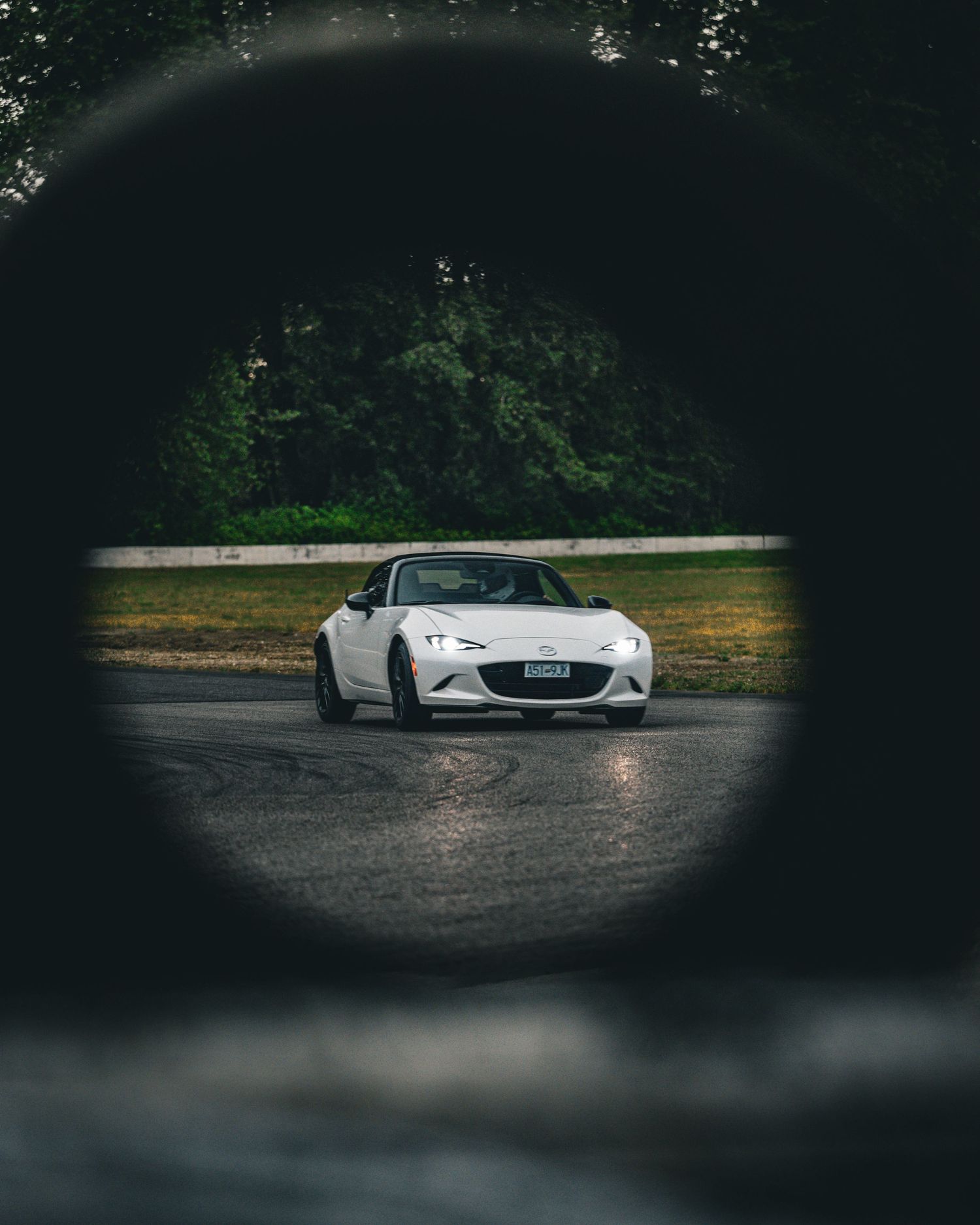 White Mazda MX-5 Miata framed through a circular opening on a wet racing track.