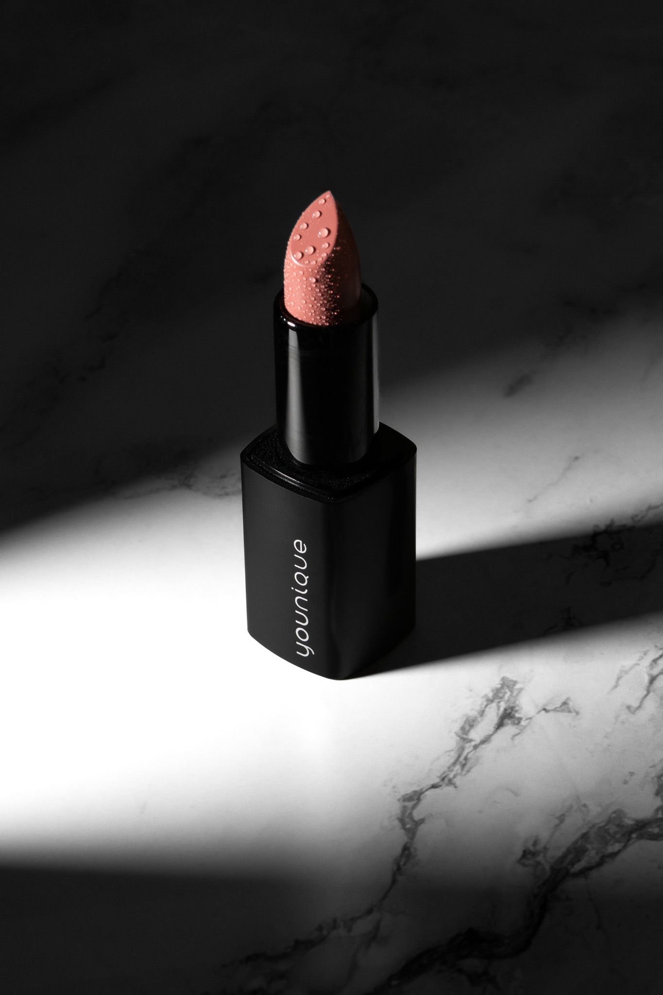 Younique pink shimmer lipstick in black tube on marble surface with dramatic shadow lighting.