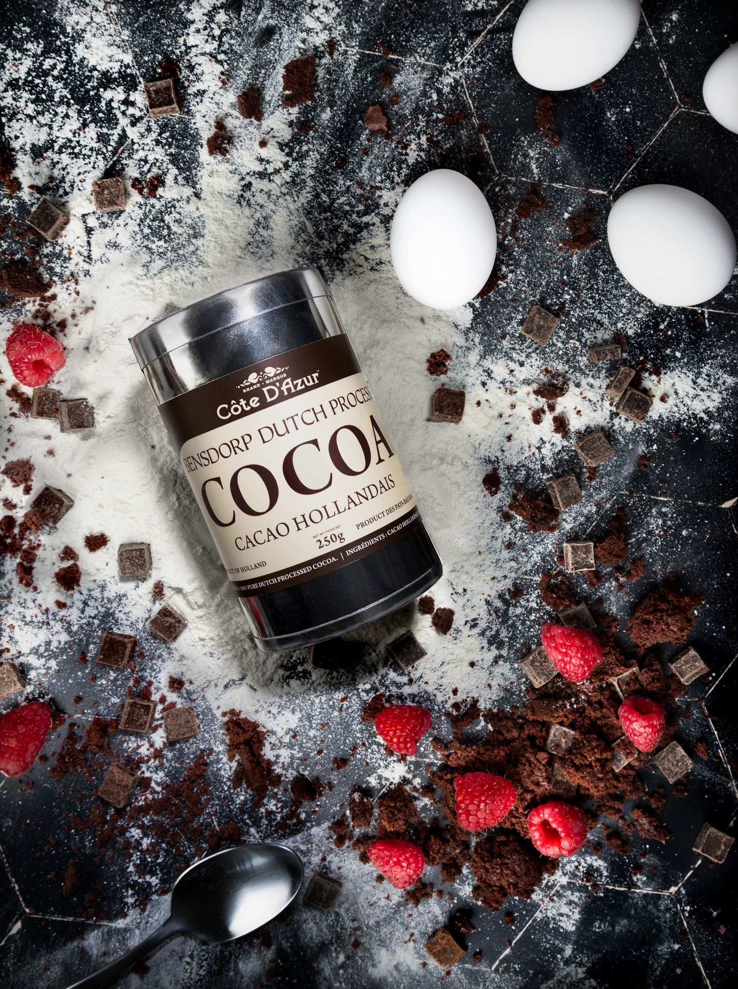 Cote D'Ivoire Dutch Process Cocoa tin surrounded by raspberries, chocolate, eggs, and flour.