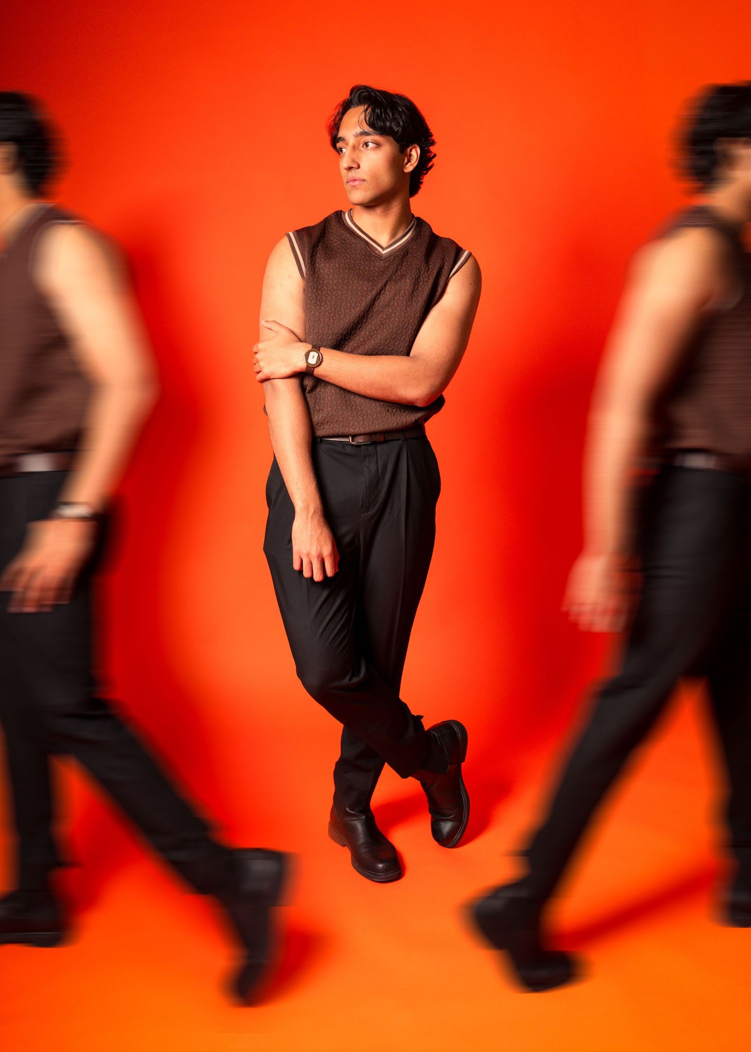 Man in brown sleeveless top and black pants poses confidently against vibrant red background with motion-blurred figures.