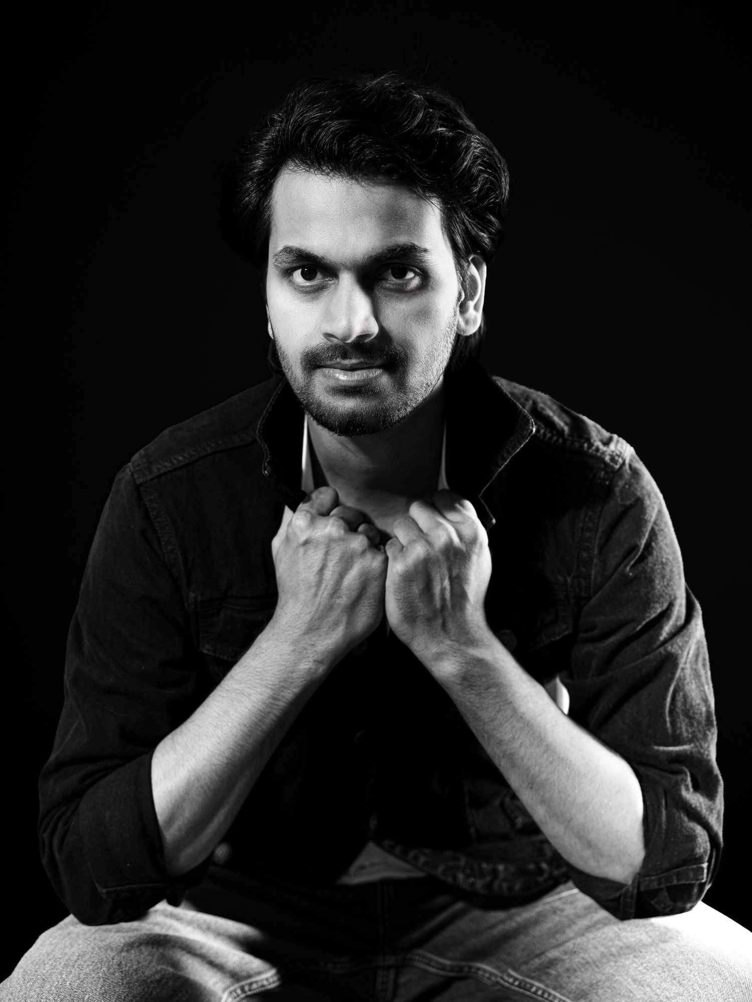 Black and white portrait of a young man with beard, hands clasped, wearing dark shirt, studio photography.