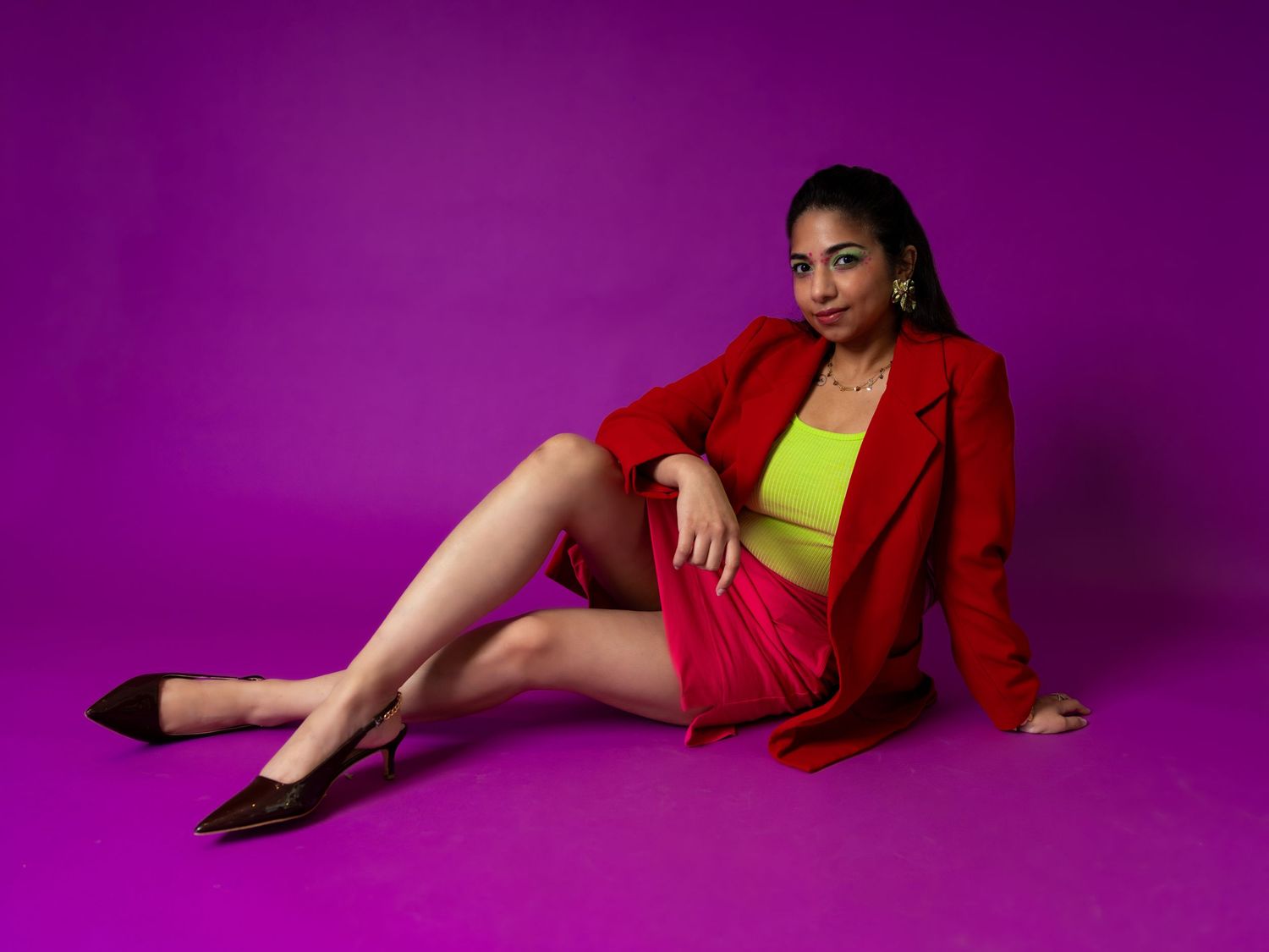 Woman in red blazer and skirt with neon yellow top poses on purple background.