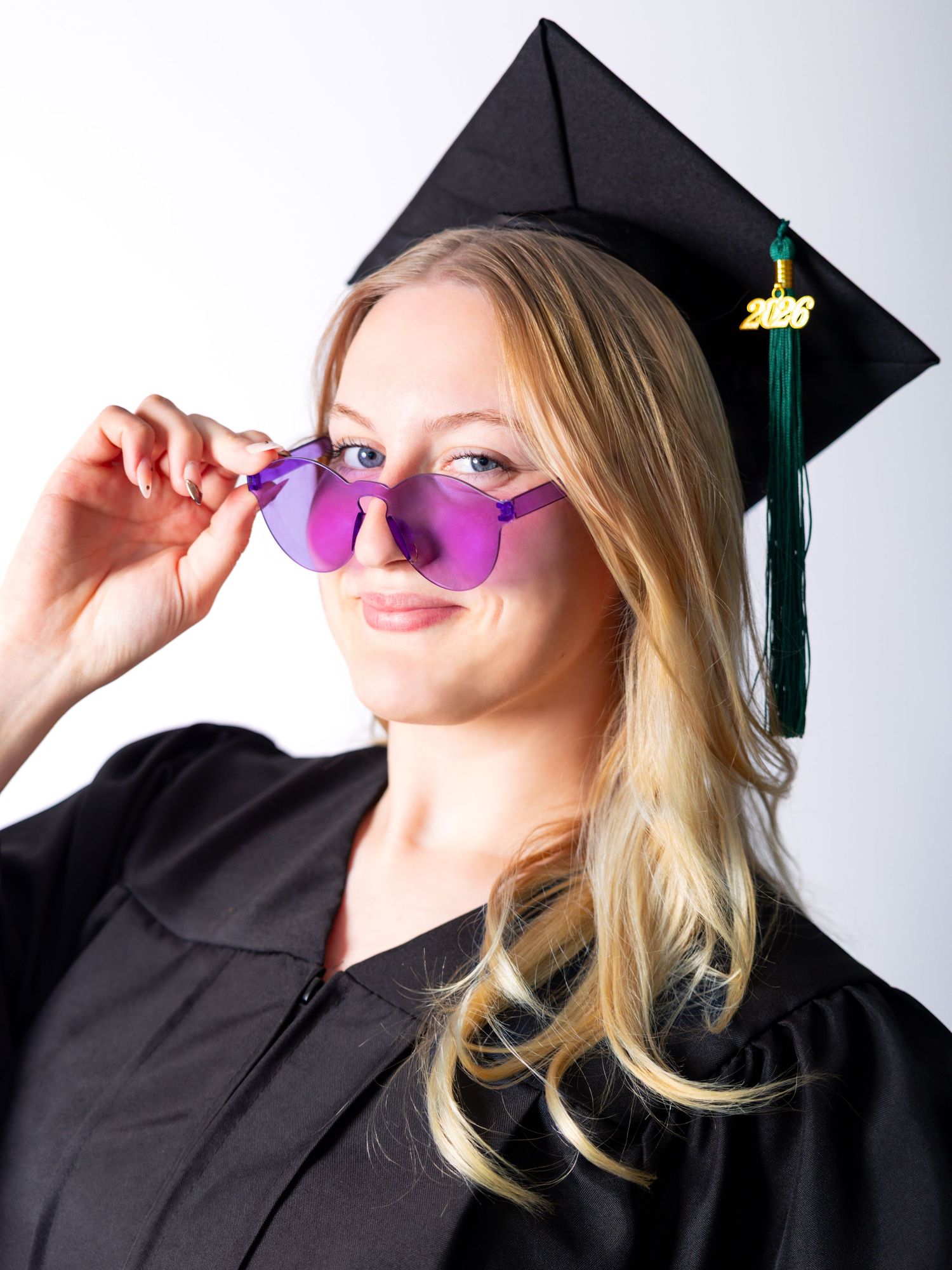 Blonde graduate in black cap and gown posing with purple tinted rimless sunglasses, green tassel, class of 2023.