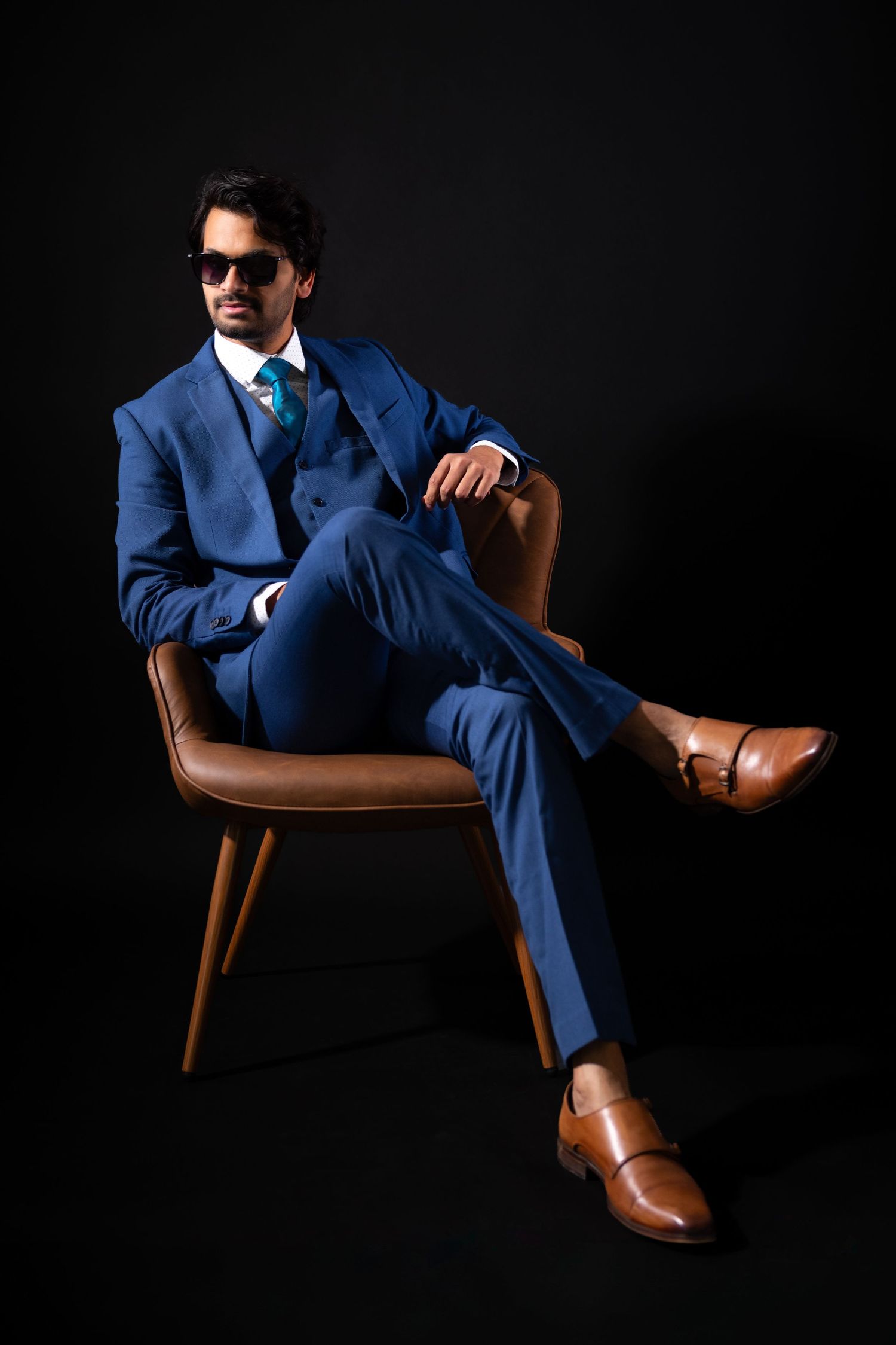 Man in blue three-piece suit, sunglasses, and brown monk strap shoes seated on leather chair against black background.