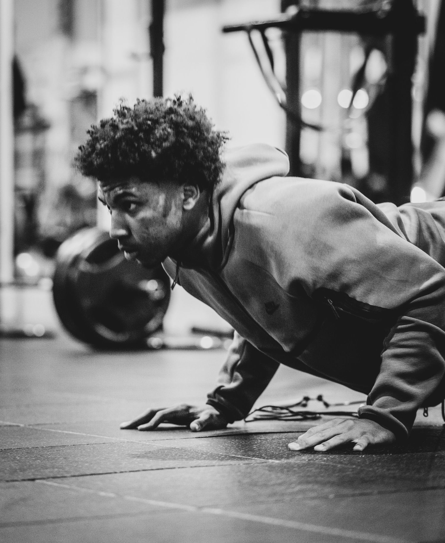 Black and white photo of a man doing push-ups in a gym, wearing a hoodie, with weights visible in the background.