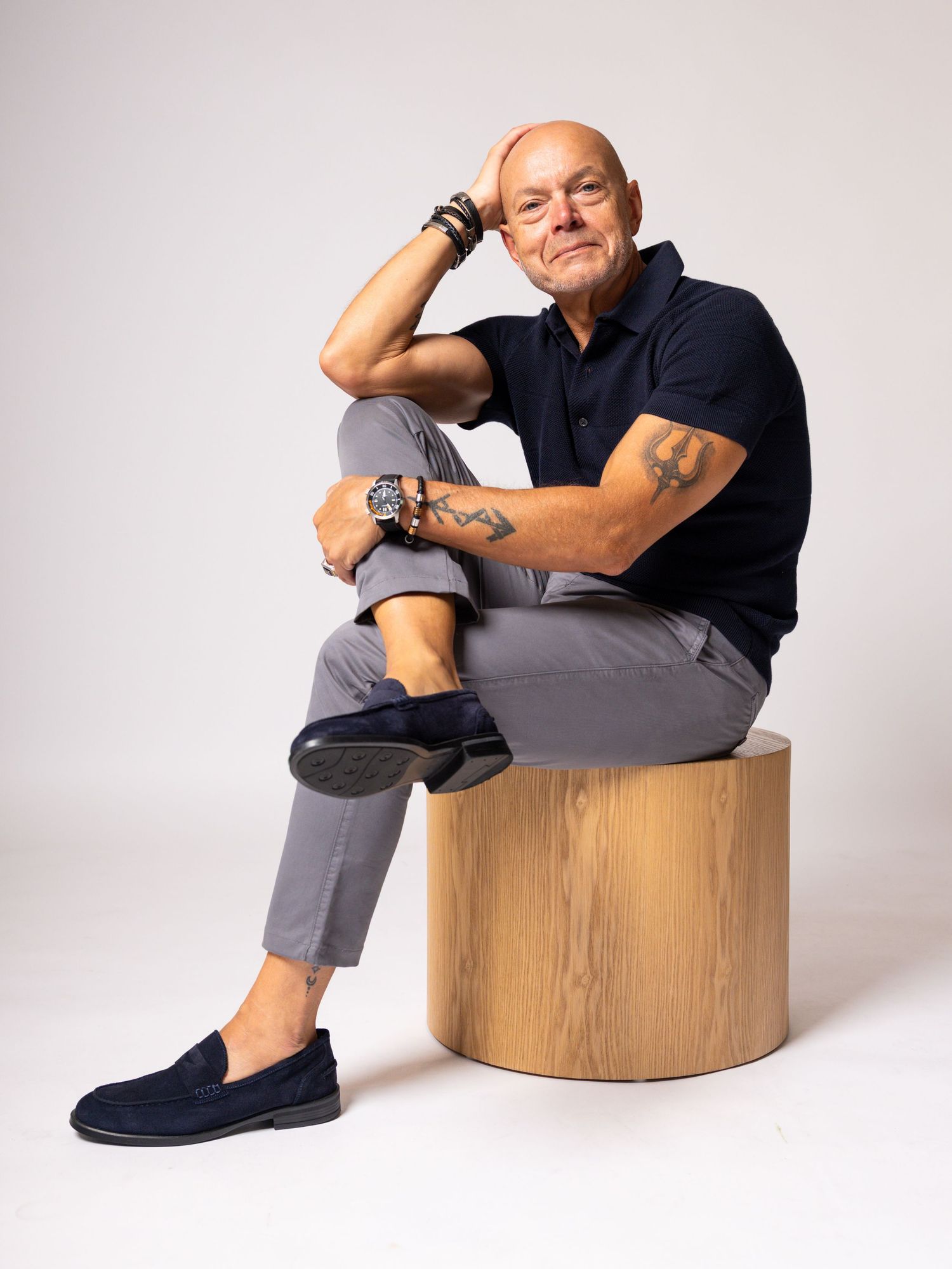 Middle-aged man with tattoos sits on wooden stool, wearing navy polo, gray pants, and blue loafers in studio setting.