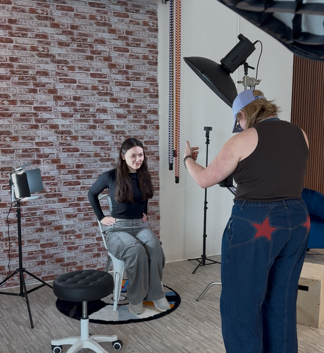 Photographer directing a female model seated on a stool in a professional photo studio with brick wall backdrop.