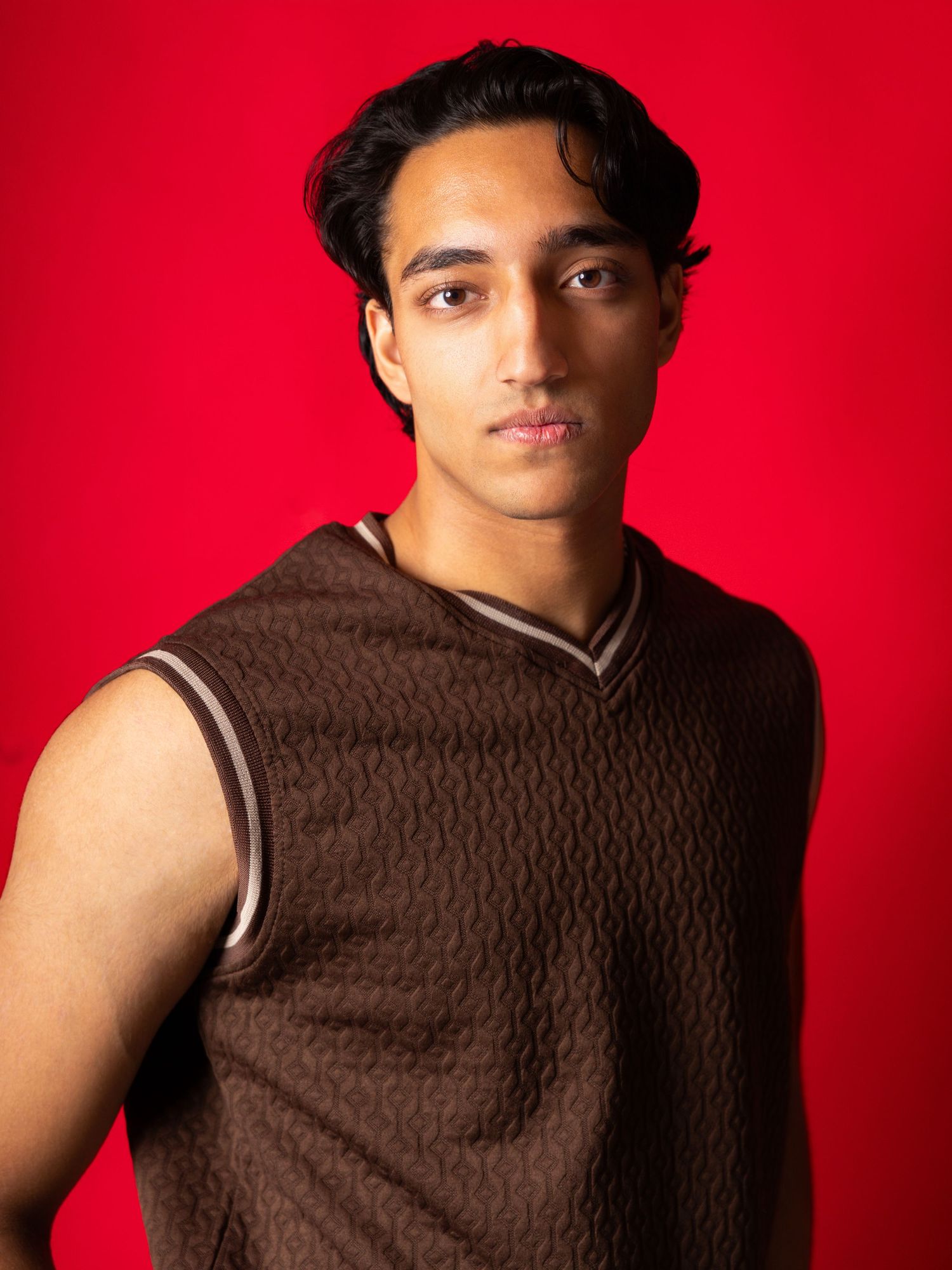 Young man in brown patterned vest posing against a bold red background in a professional studio portrait.