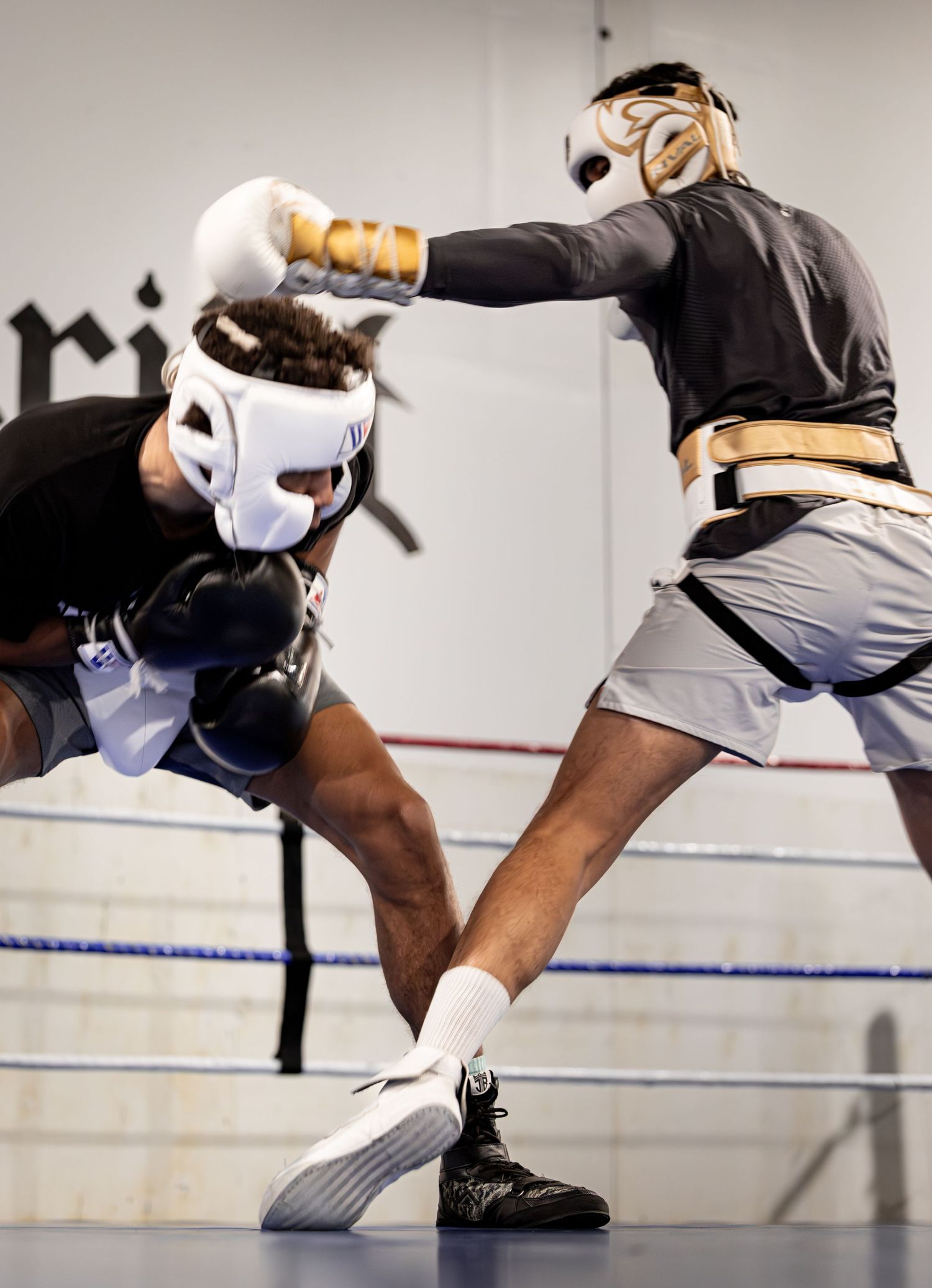 Two boxers sparring in a boxing ring, one throwing a punch while wearing headgear and white gloves.