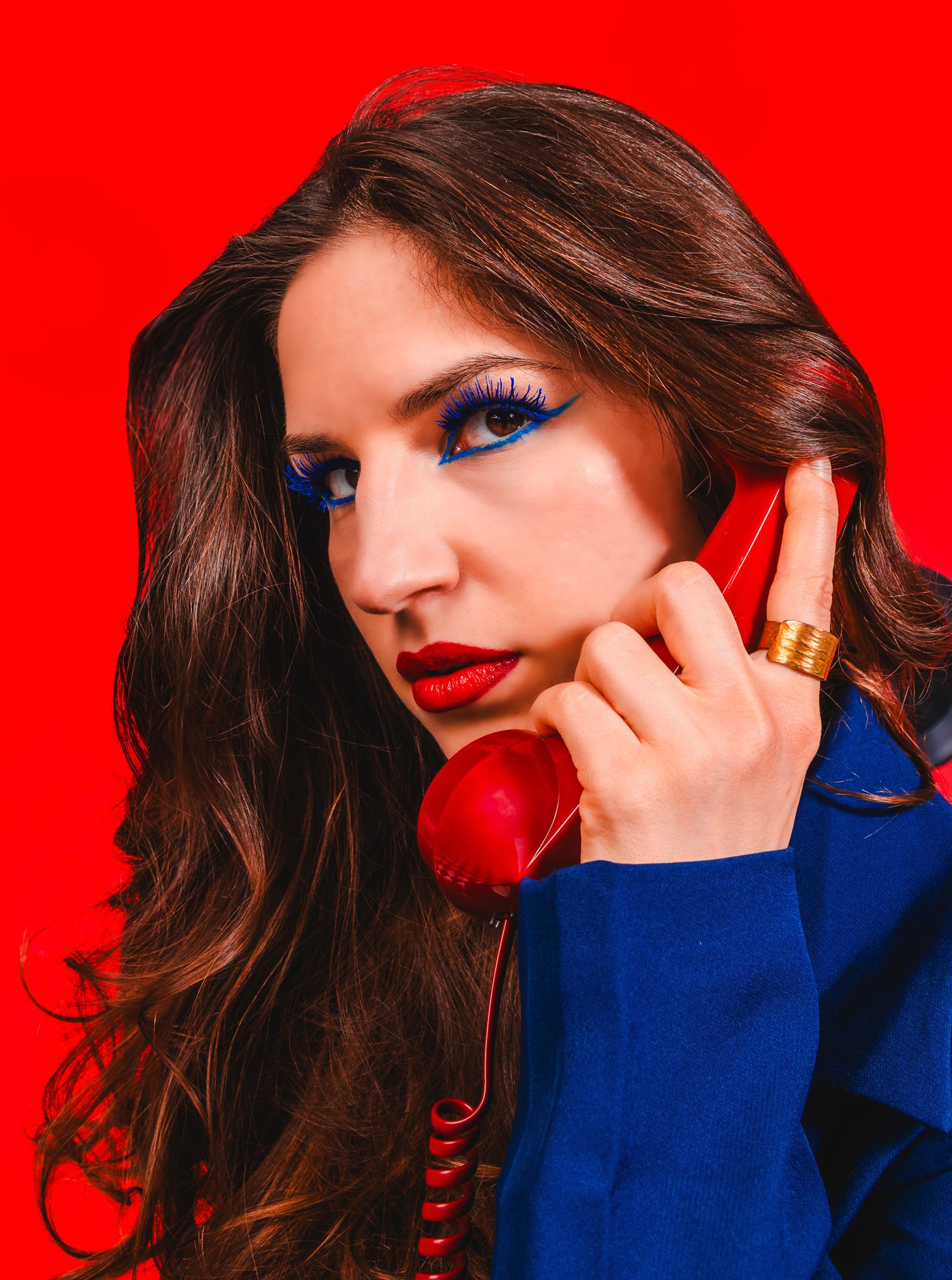 Stylish brunette woman holding a red retro phone with bold blue eye makeup on red background.