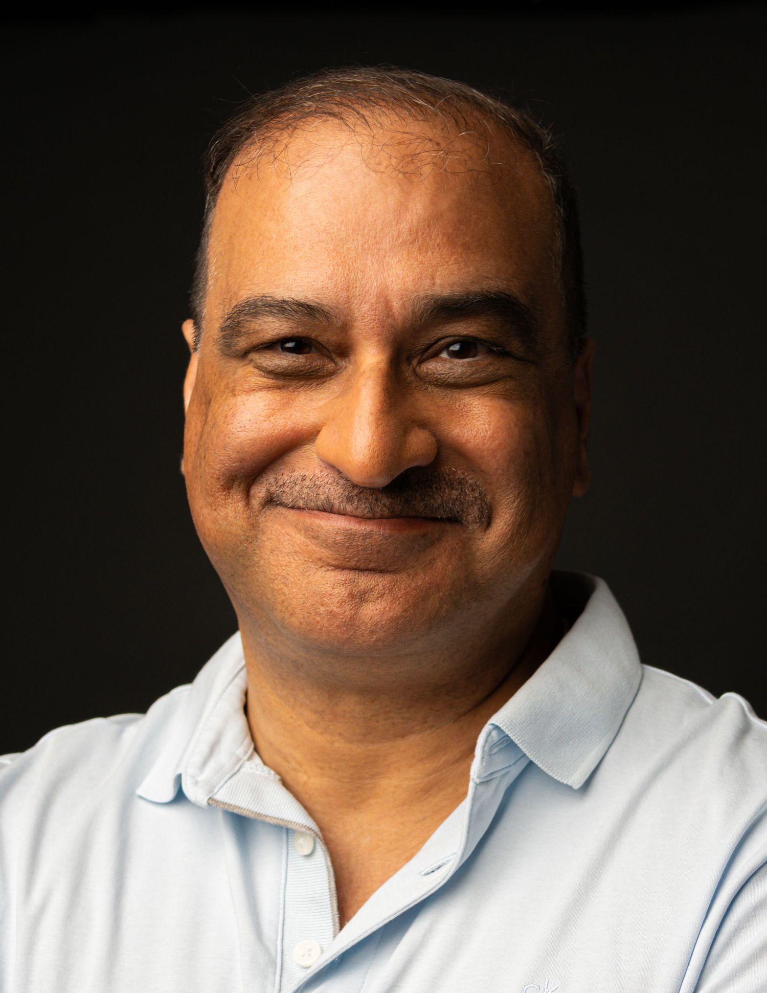 Portrait of a cheerful middle-aged man in a light blue collared shirt smiling against a dark backdrop.