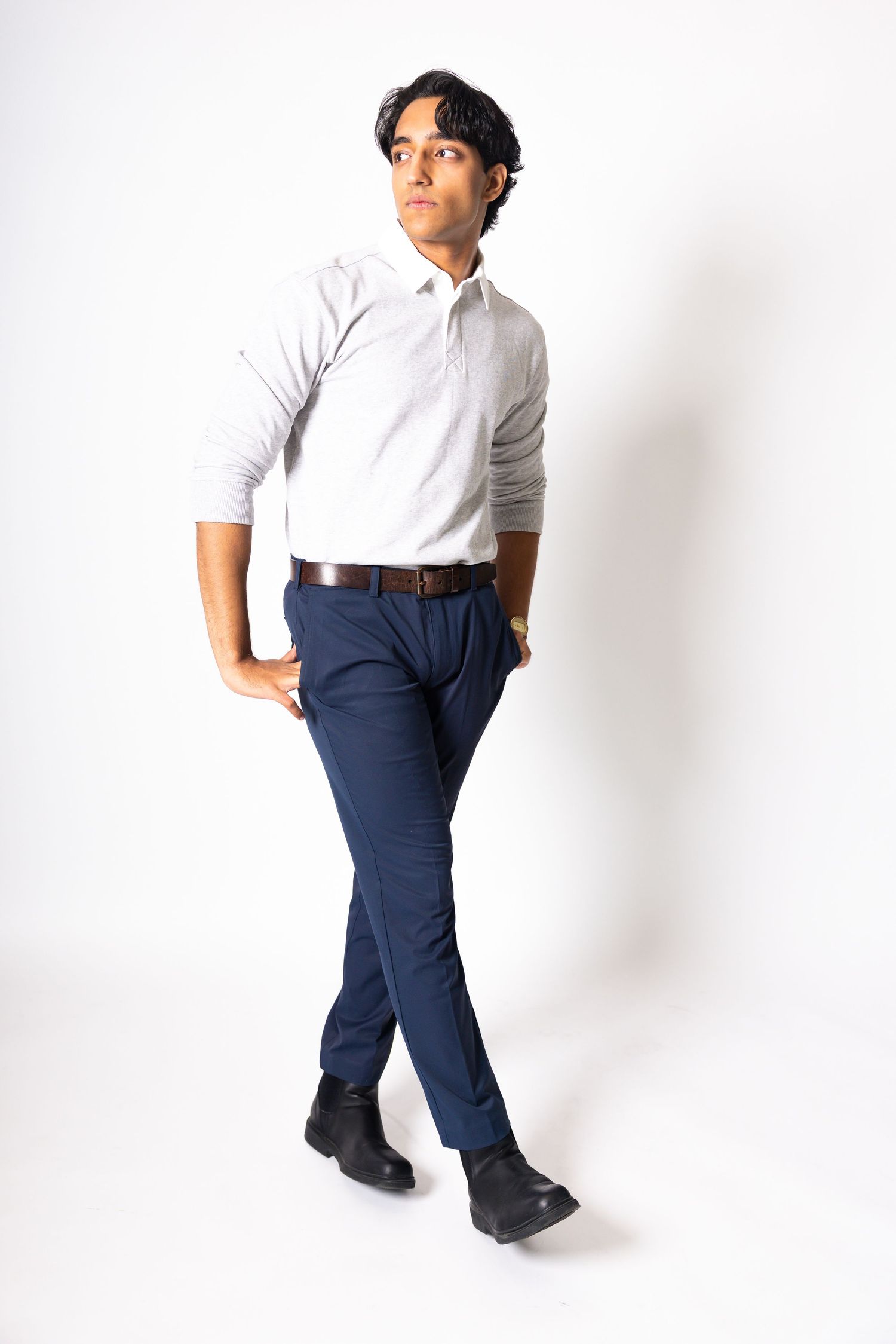 Young man in white polo shirt, navy trousers, and black shoes poses in a full-length studio portrait.