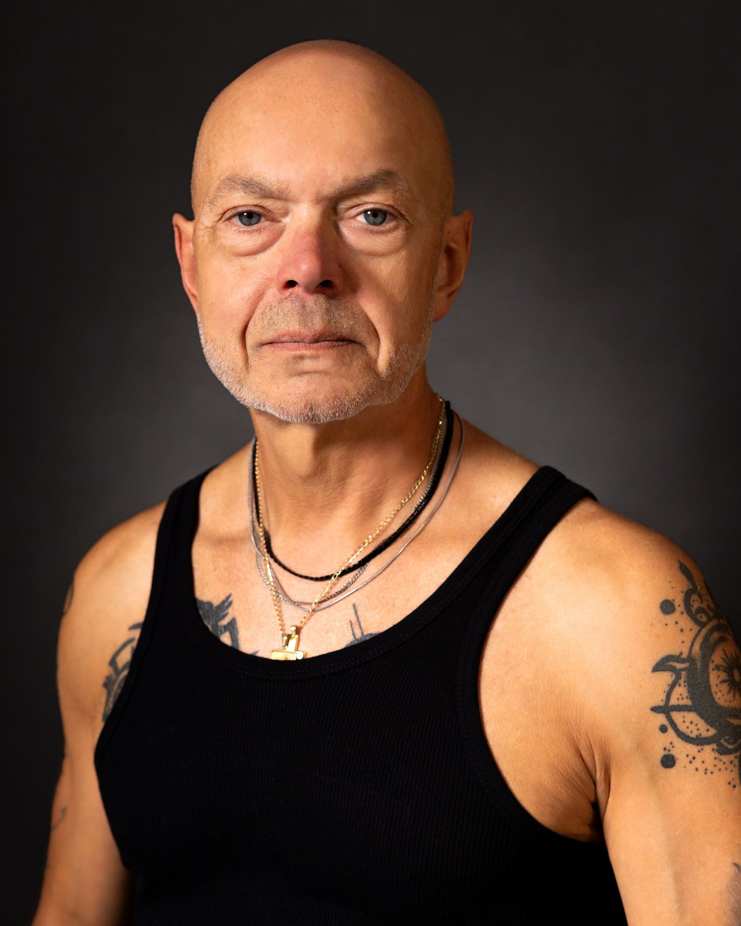Bald man with tattoos wearing a black tank top and gold necklace poses for a professional portrait photo.