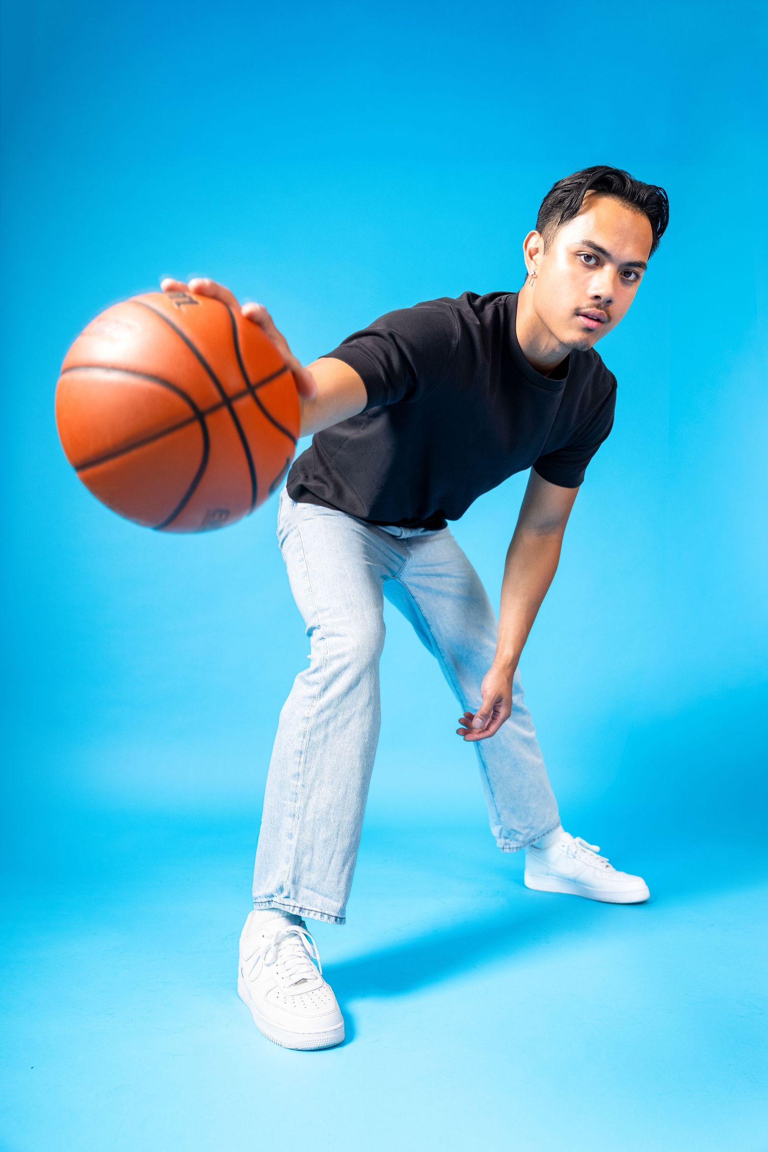 Young man in black t-shirt and jeans dribbling a basketball against a bright blue background.