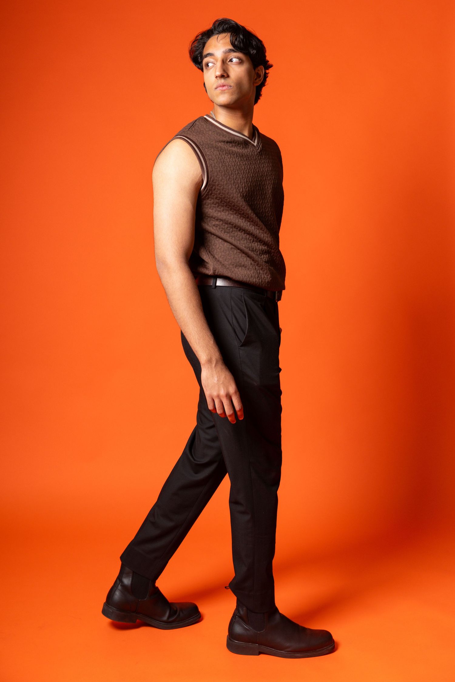 Young man in brown knit vest and black trousers posing confidently against vibrant orange background.