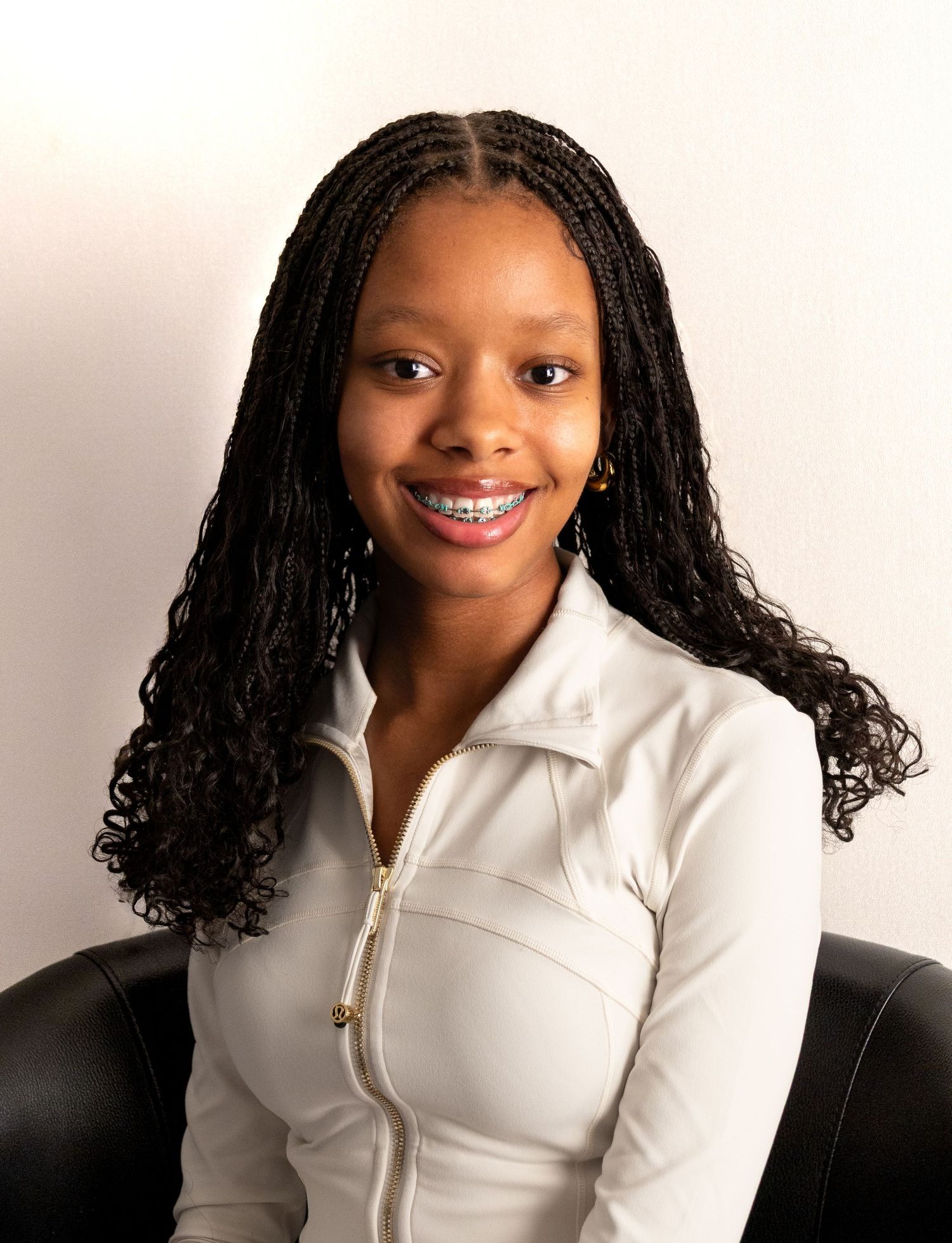 Smiling young woman with box braids and braces wears a white zip-up jacket in a professional portrait.