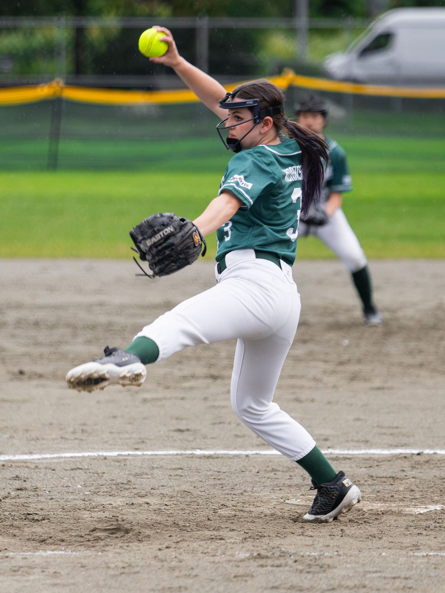 Female softball pitcher in green uniform mid-windup, releasing yellow ball during competitive game on dirt field.