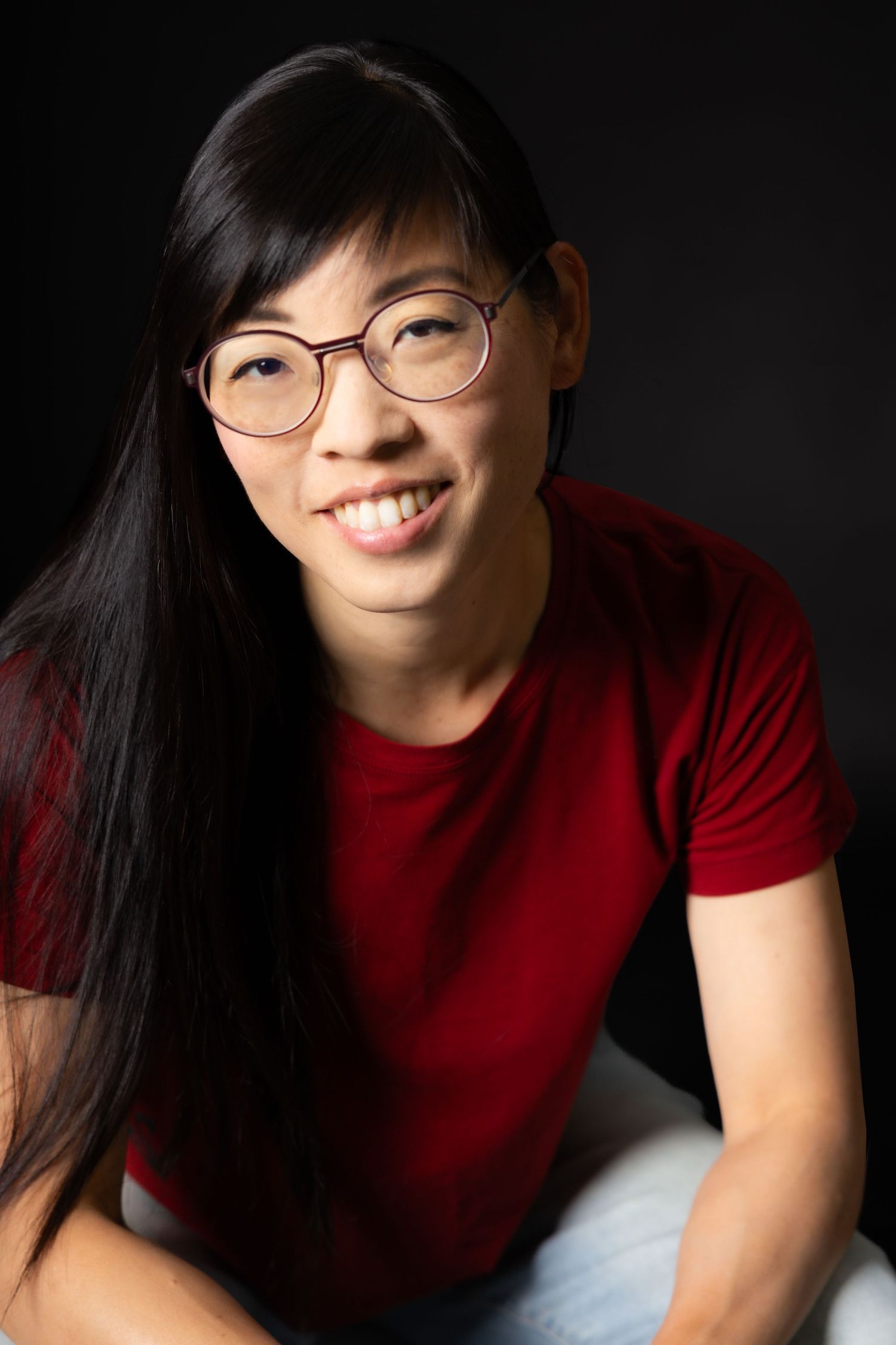 Smiling Asian woman with long black hair and round glasses wearing a red t-shirt against a dark background.