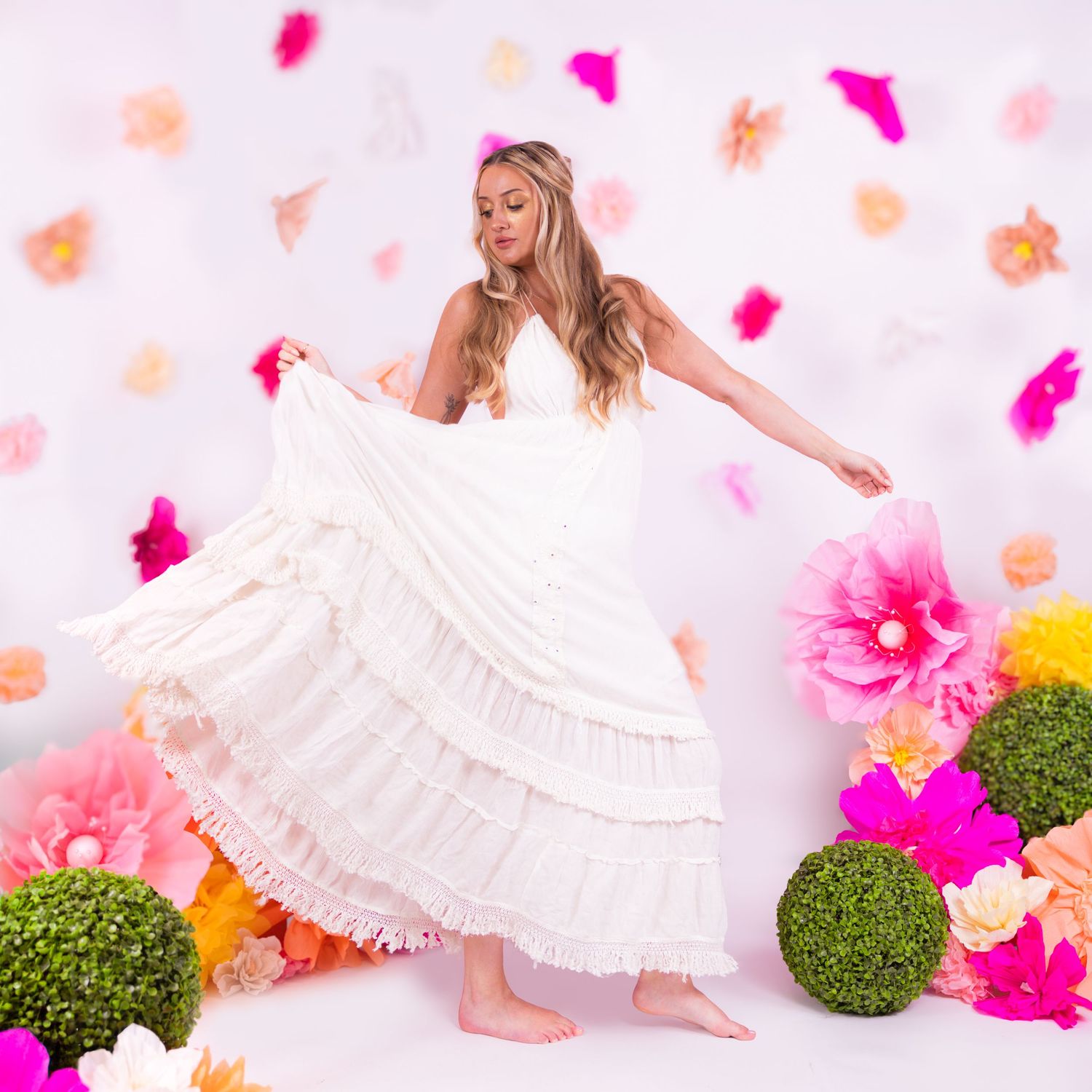 Woman in white ruffled wedding gown twirling among colorful paper flowers and topiary balls on white backdrop.