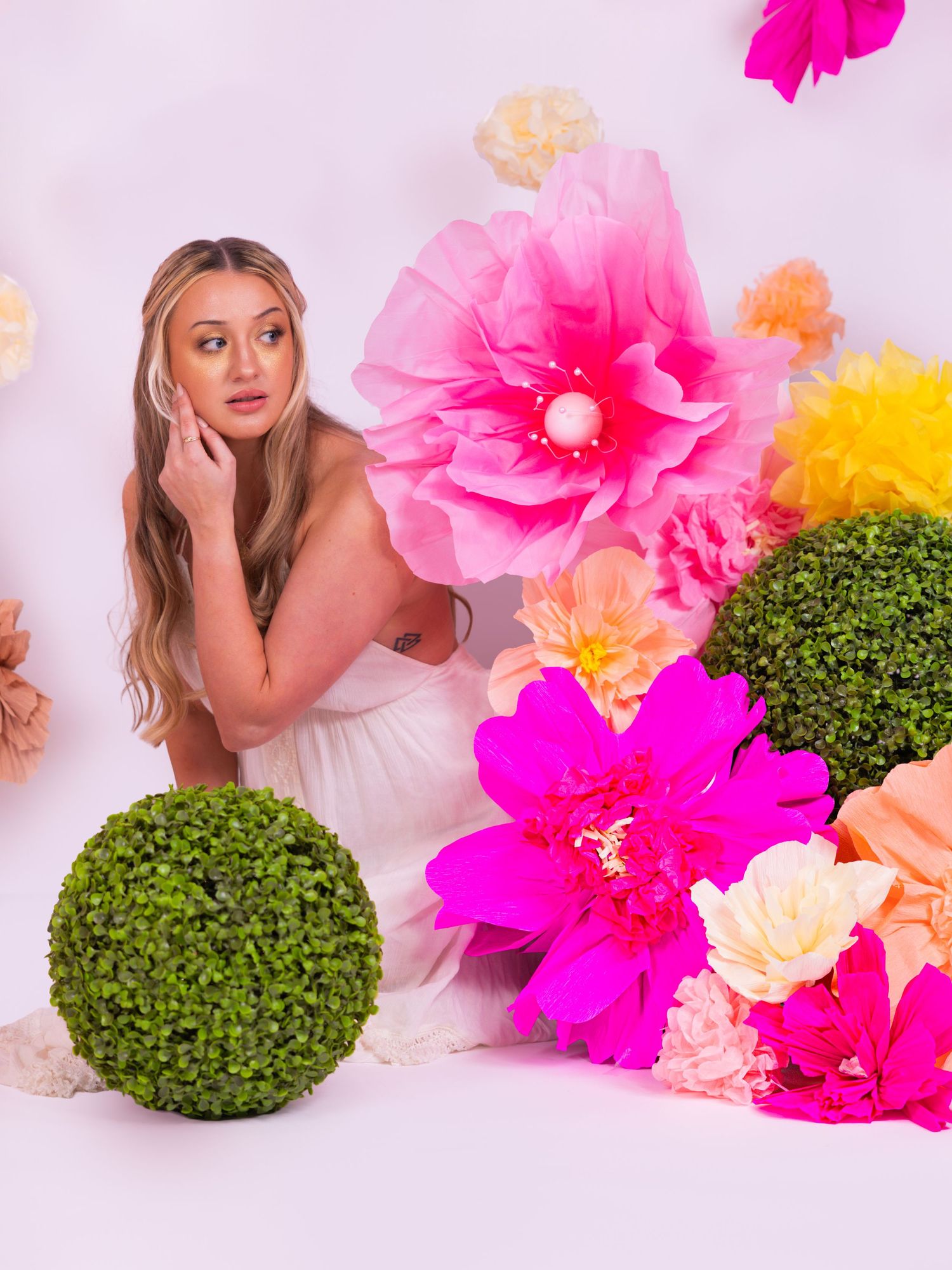 Woman in white dress posing with colorful giant paper flowers and green topiary balls on white background.