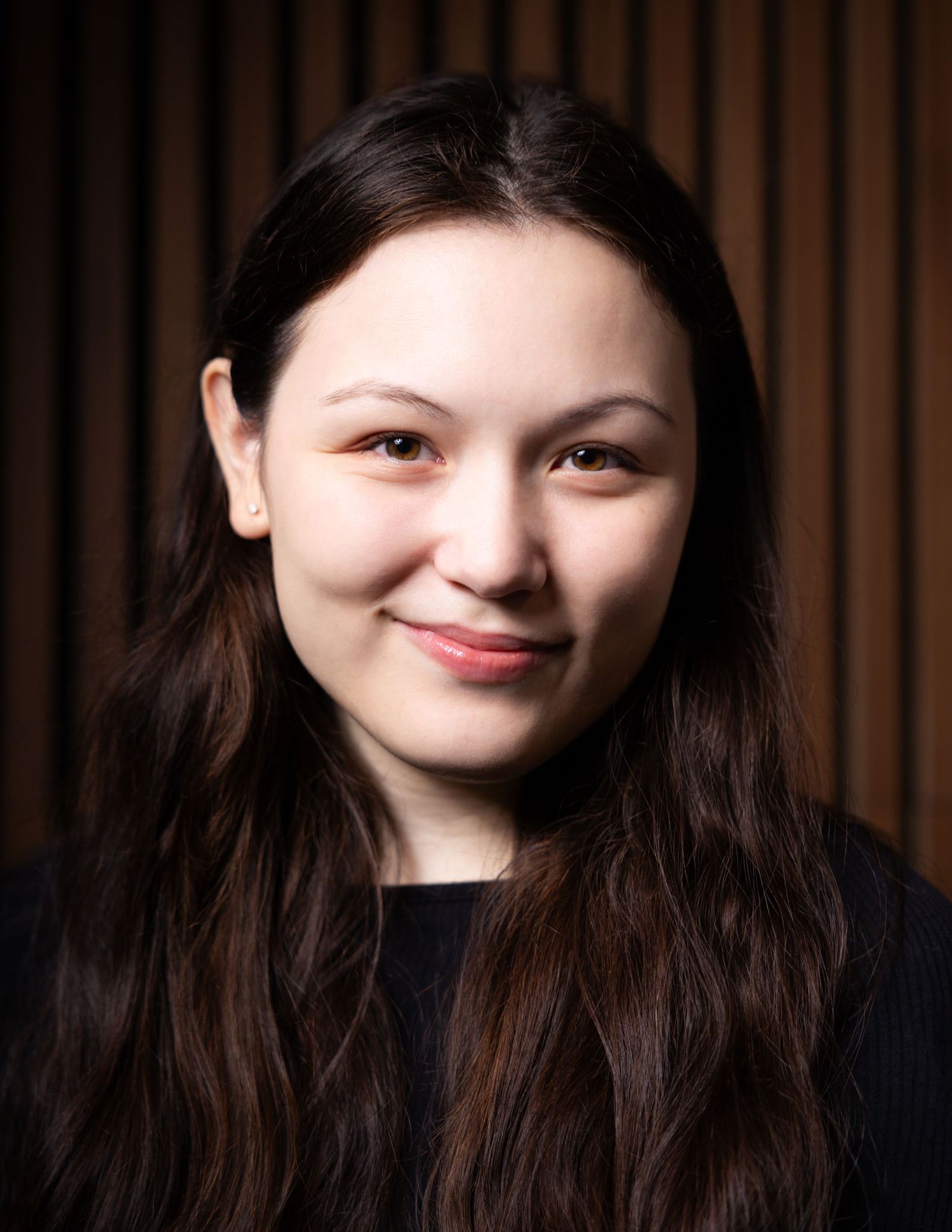 Young woman with long dark hair smiling warmly, wearing a dark top against a wooden panel background.