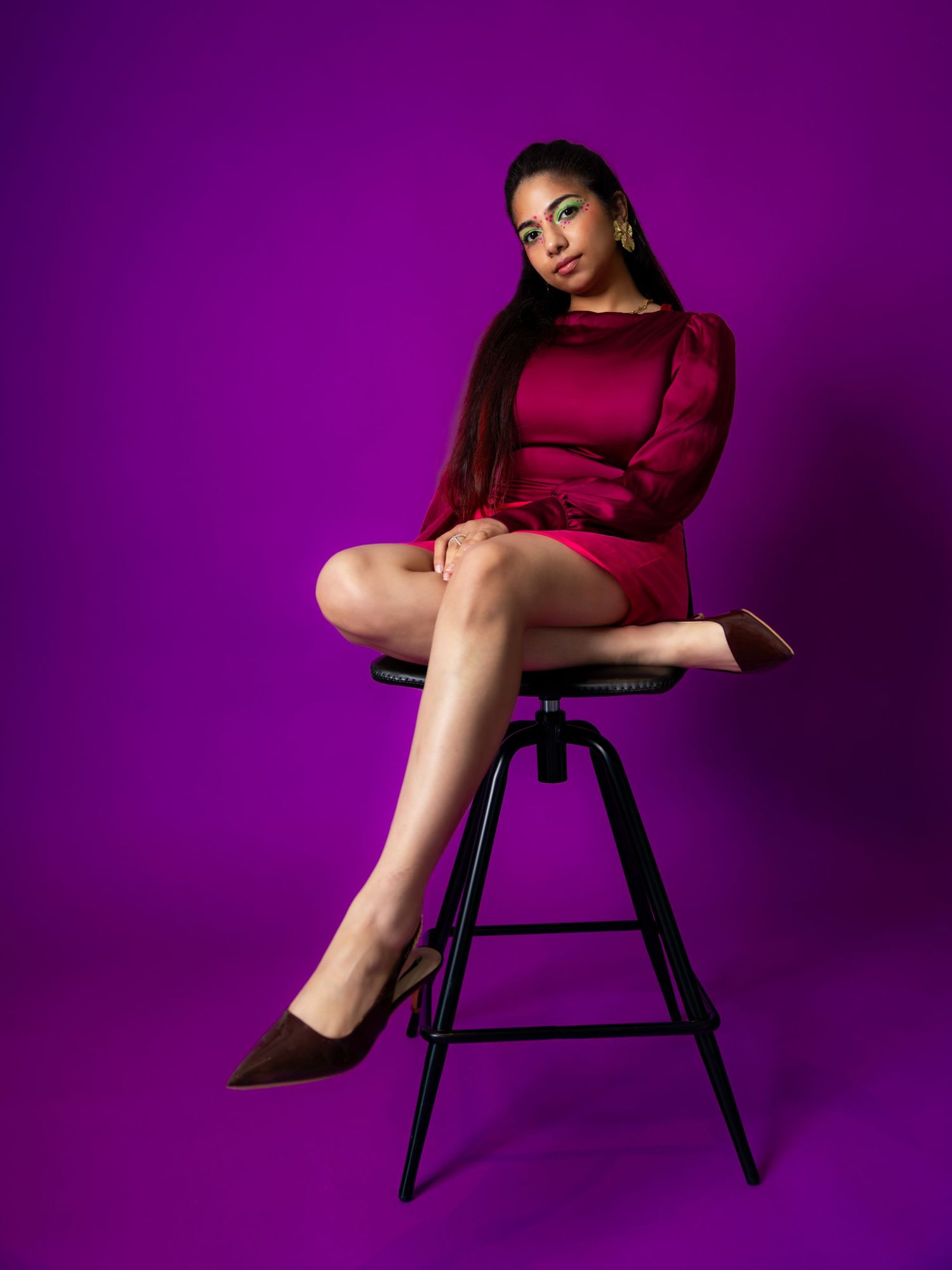 A young woman in a red mini dress and heels sits elegantly on a black stool against a vibrant purple background.