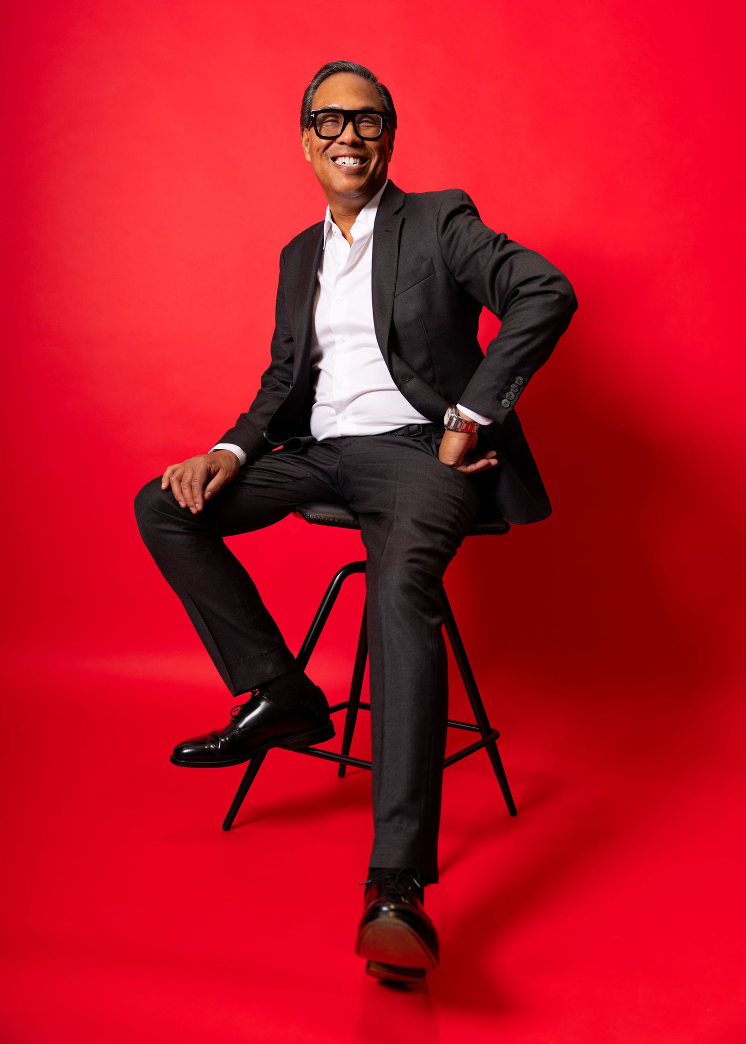 A confident person in a dark suit sits on a stool against a bold red background, smiling with crossed legs.