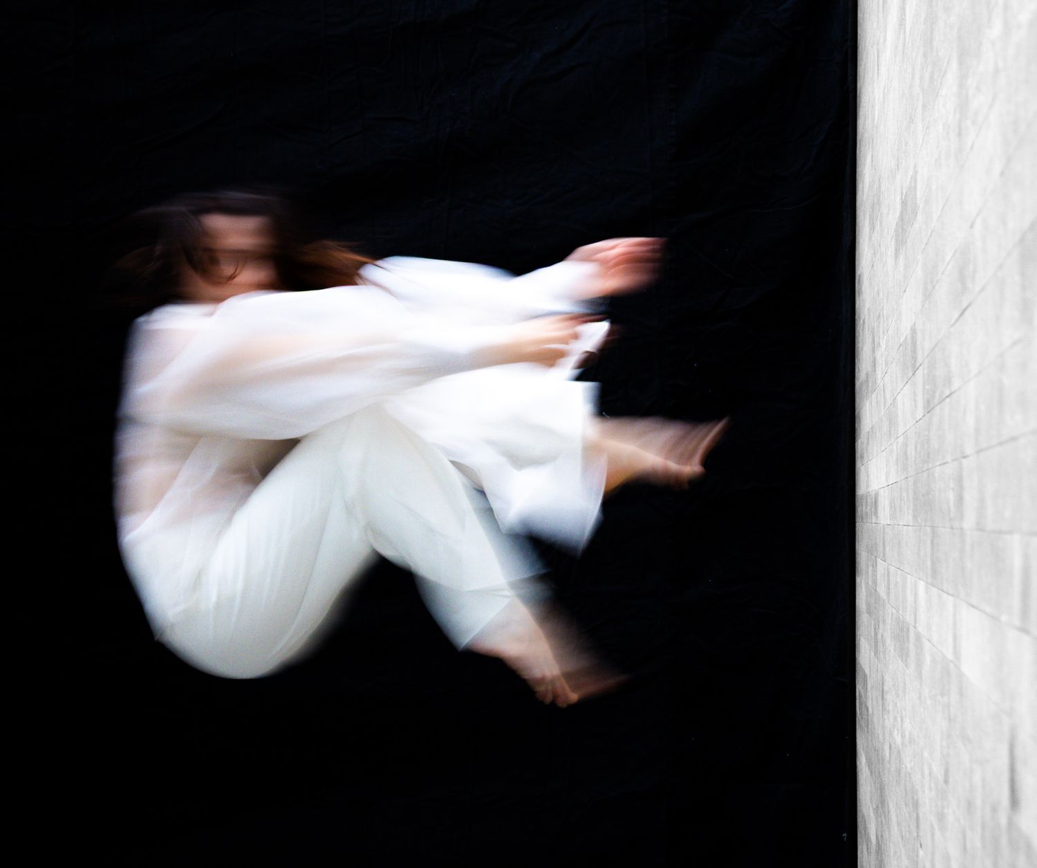 Blurred motion art photo of a dancer in white clothing curled in mid-air against a black background.