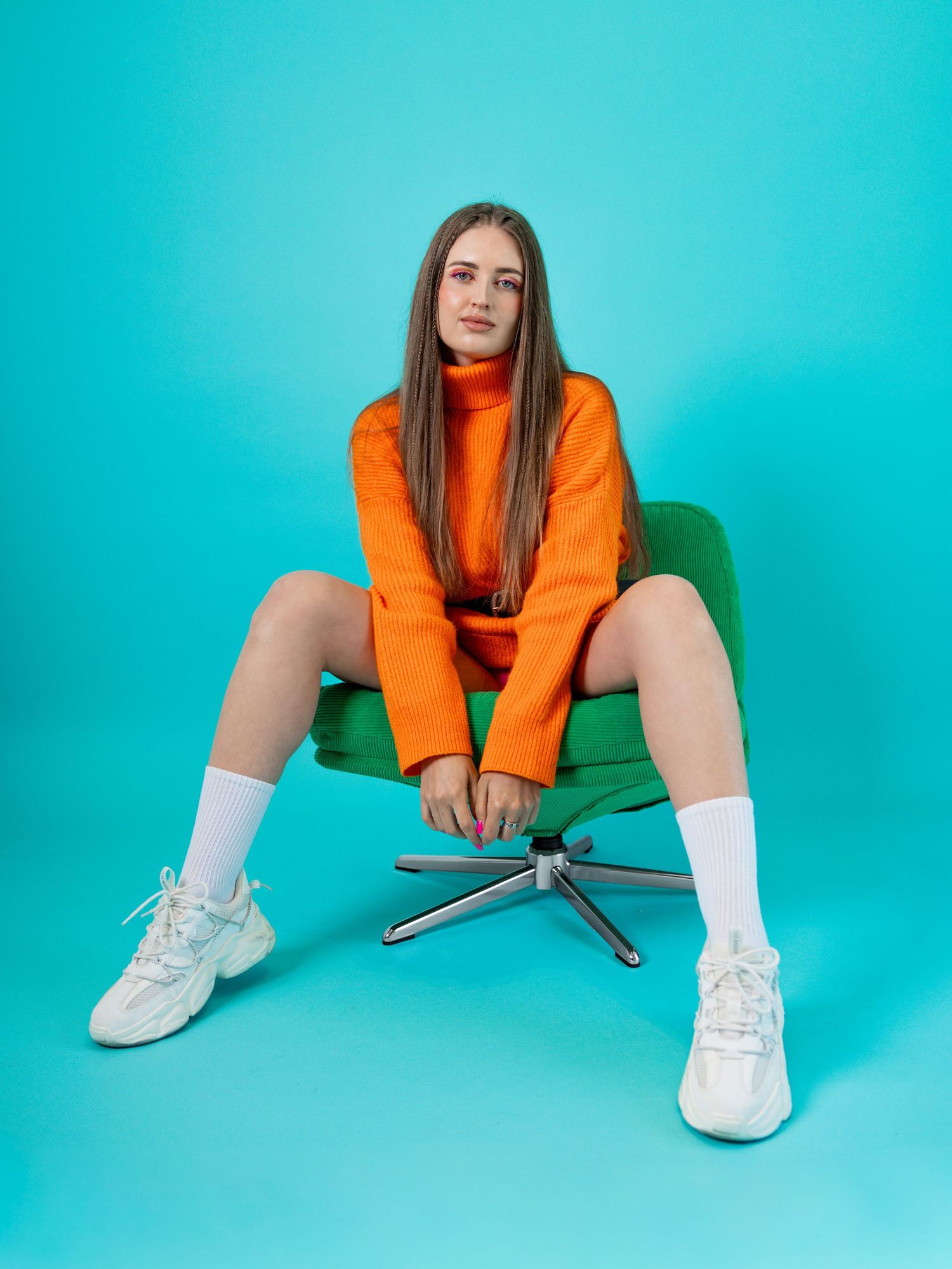 Young woman in orange turtleneck outfit sitting on green chair against teal background wearing white sneakers.