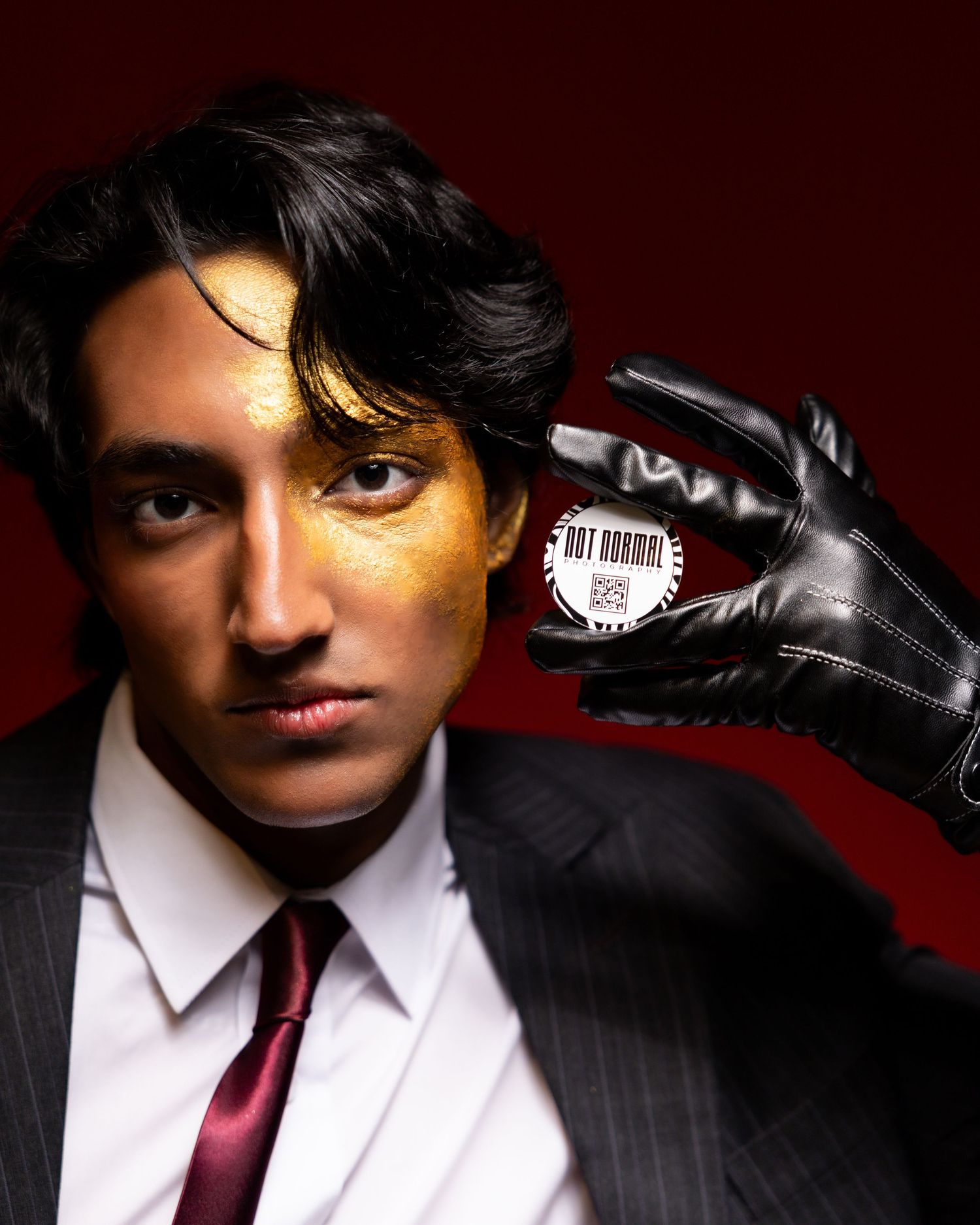 Man in suit with gold face paint holds a 'Not Normal' button while wearing a black leather glove.