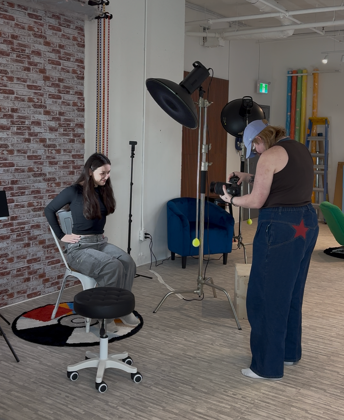 Photographer taking portrait of woman seated in studio chair with professional lighting equipment and brick wall backdrop.