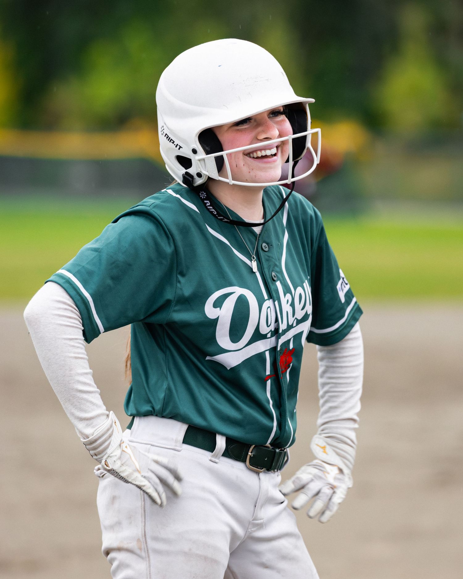 Smiling softball player in green Dakers jersey and white helmet runs bases during a game.