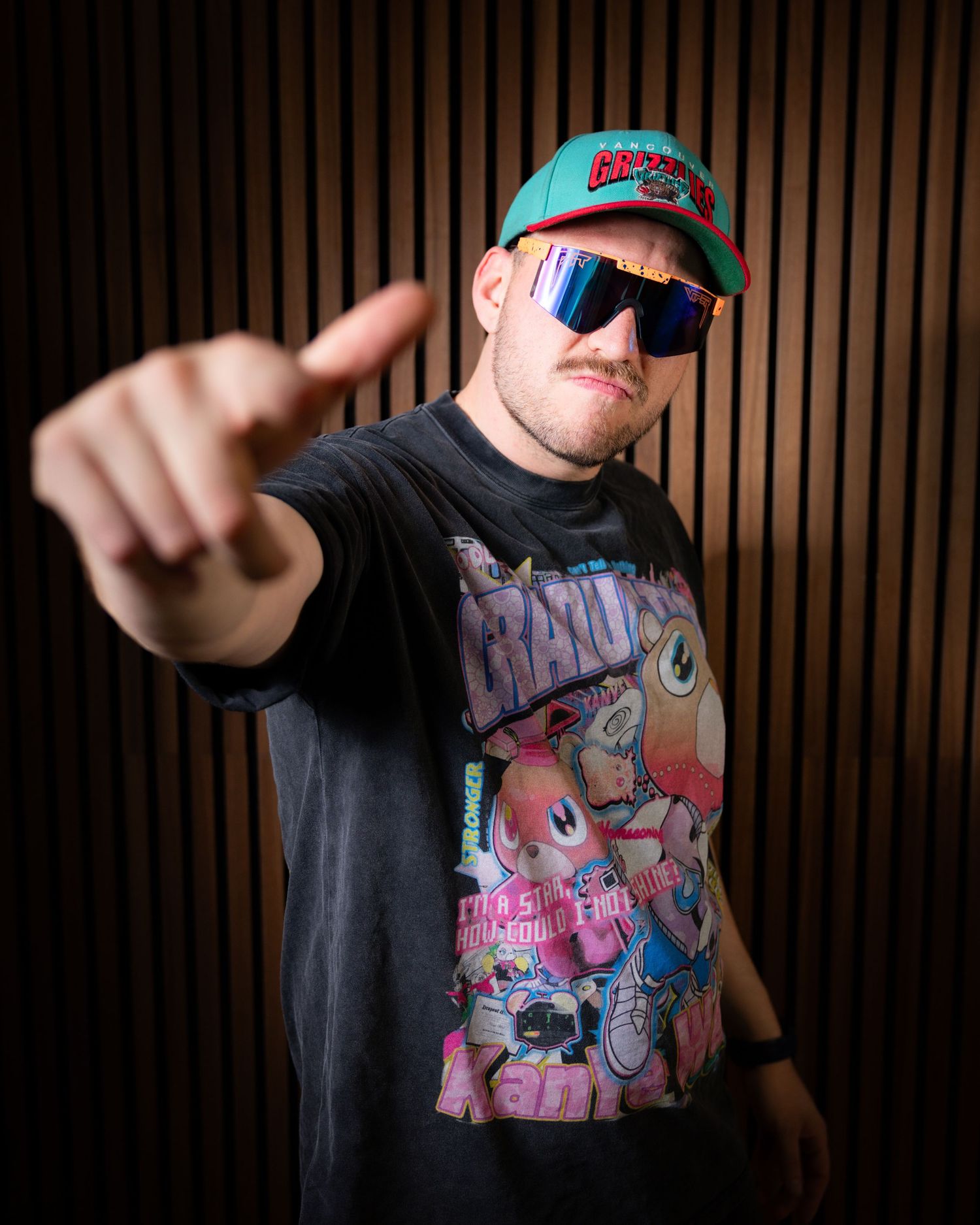 Man in graphic tee and teal cap with sunglasses pointing at camera against wooden slat wall.