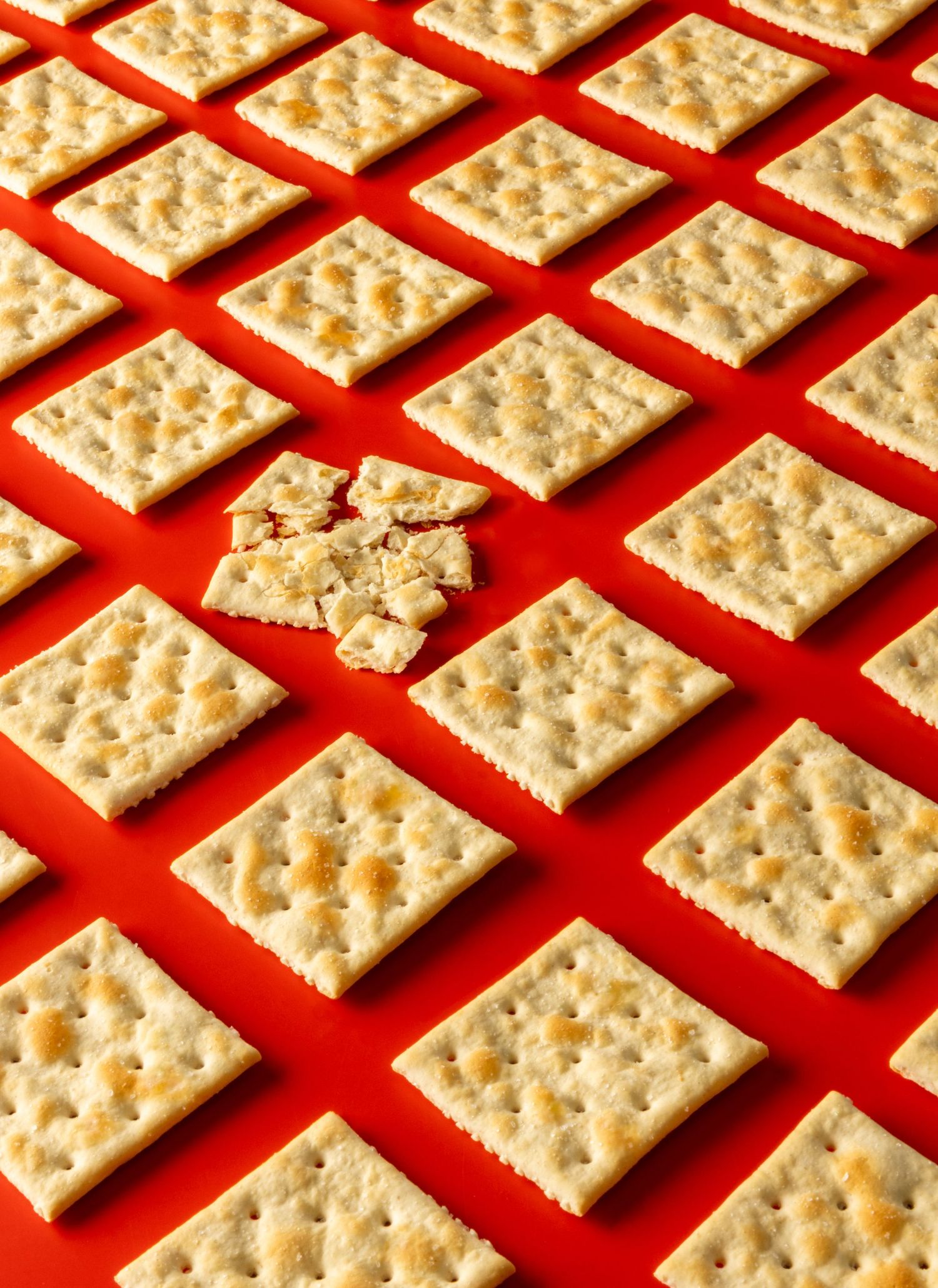 Grid pattern of square saltine crackers on red background with one cracker broken in the center.
