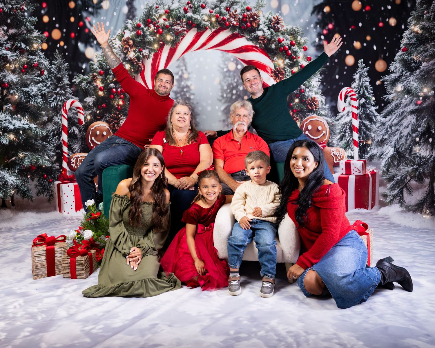 Large family posing together in festive Christmas photo with decorated trees, candy canes, and wrapped gifts in snowy scene.