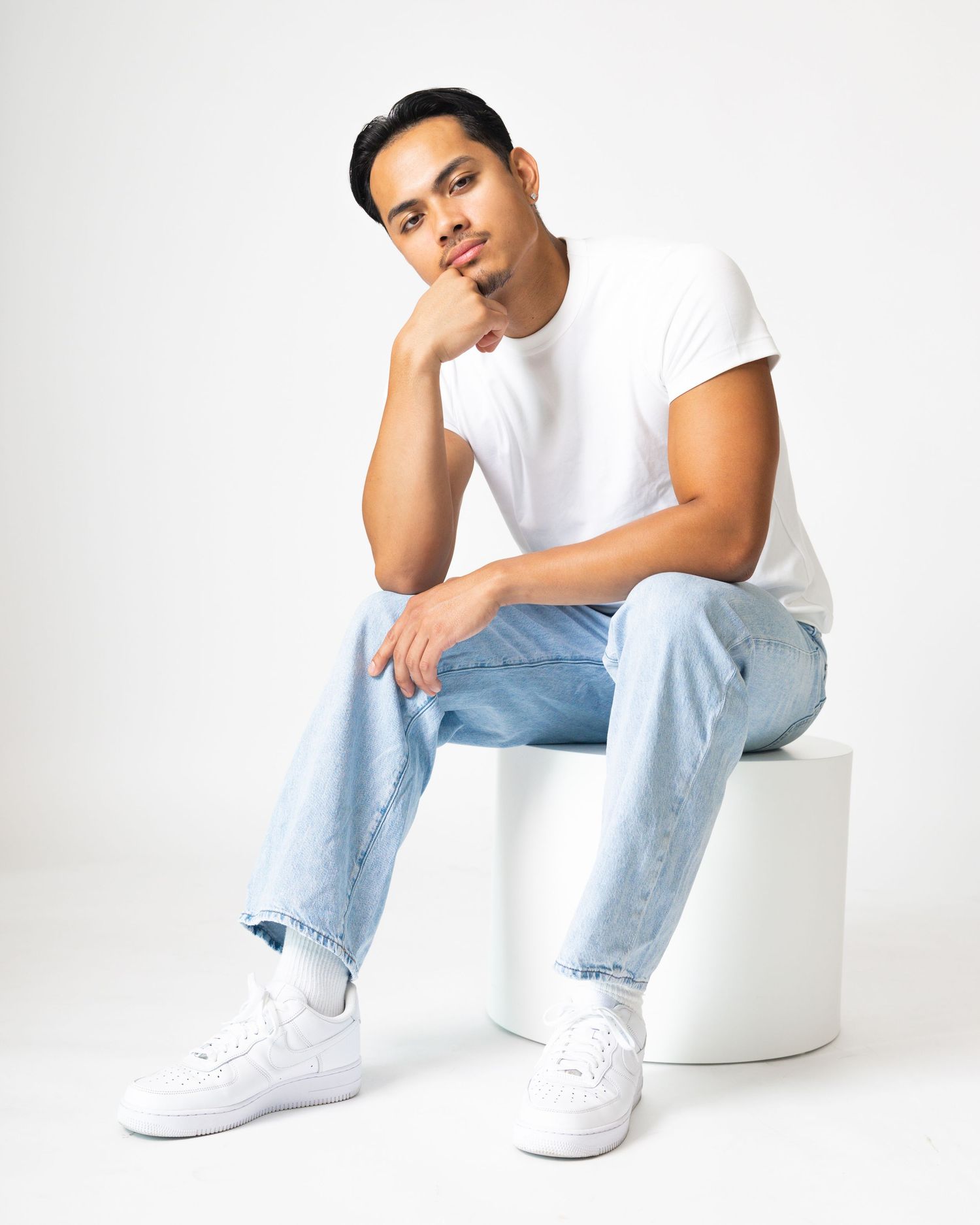 Young man posing on white cube in white t-shirt, light blue jeans, and white sneakers with hand on chin.