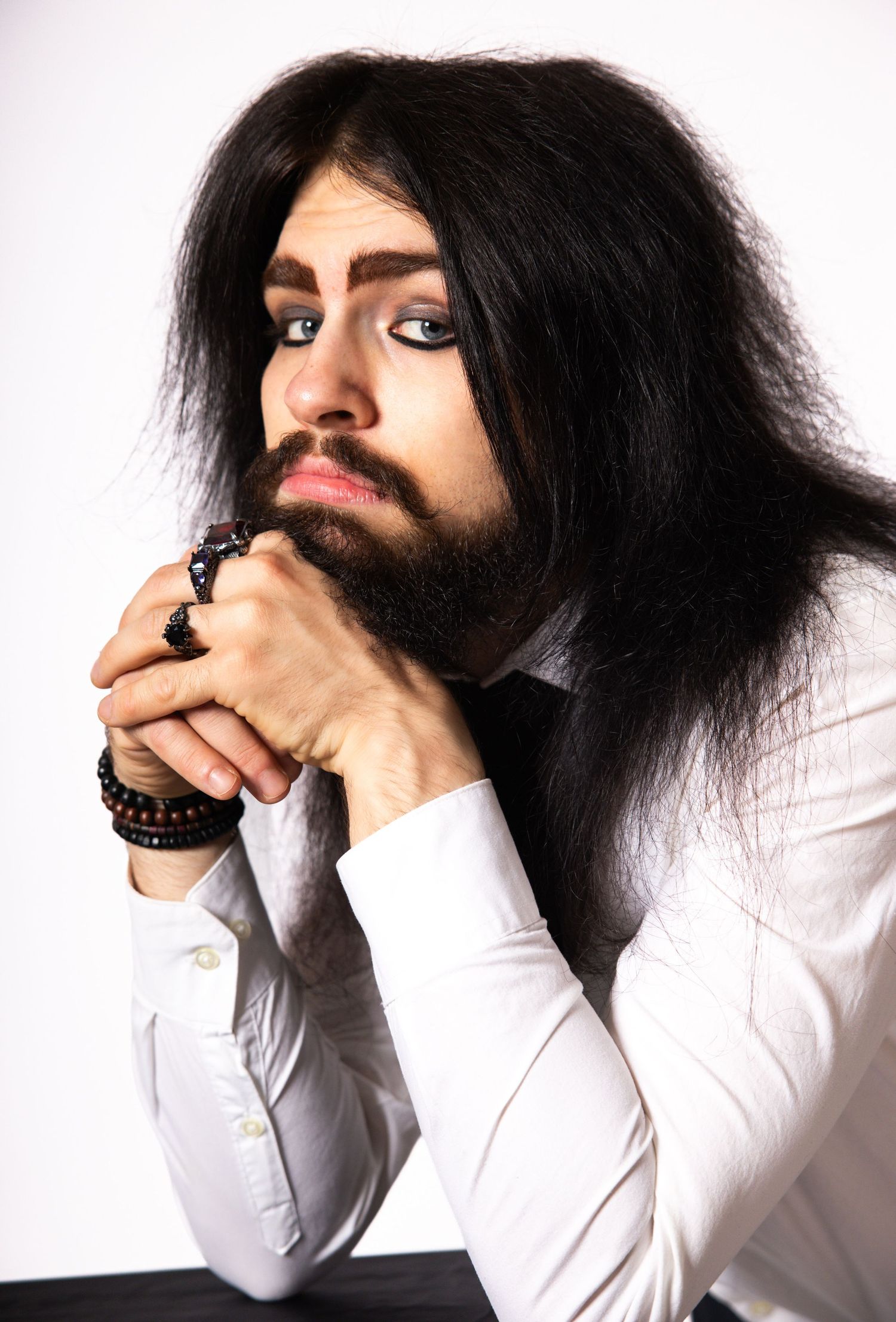 Bearded man with long black hair in white shirt, hands clasped thoughtfully, wearing beaded bracelet, studio portrait.