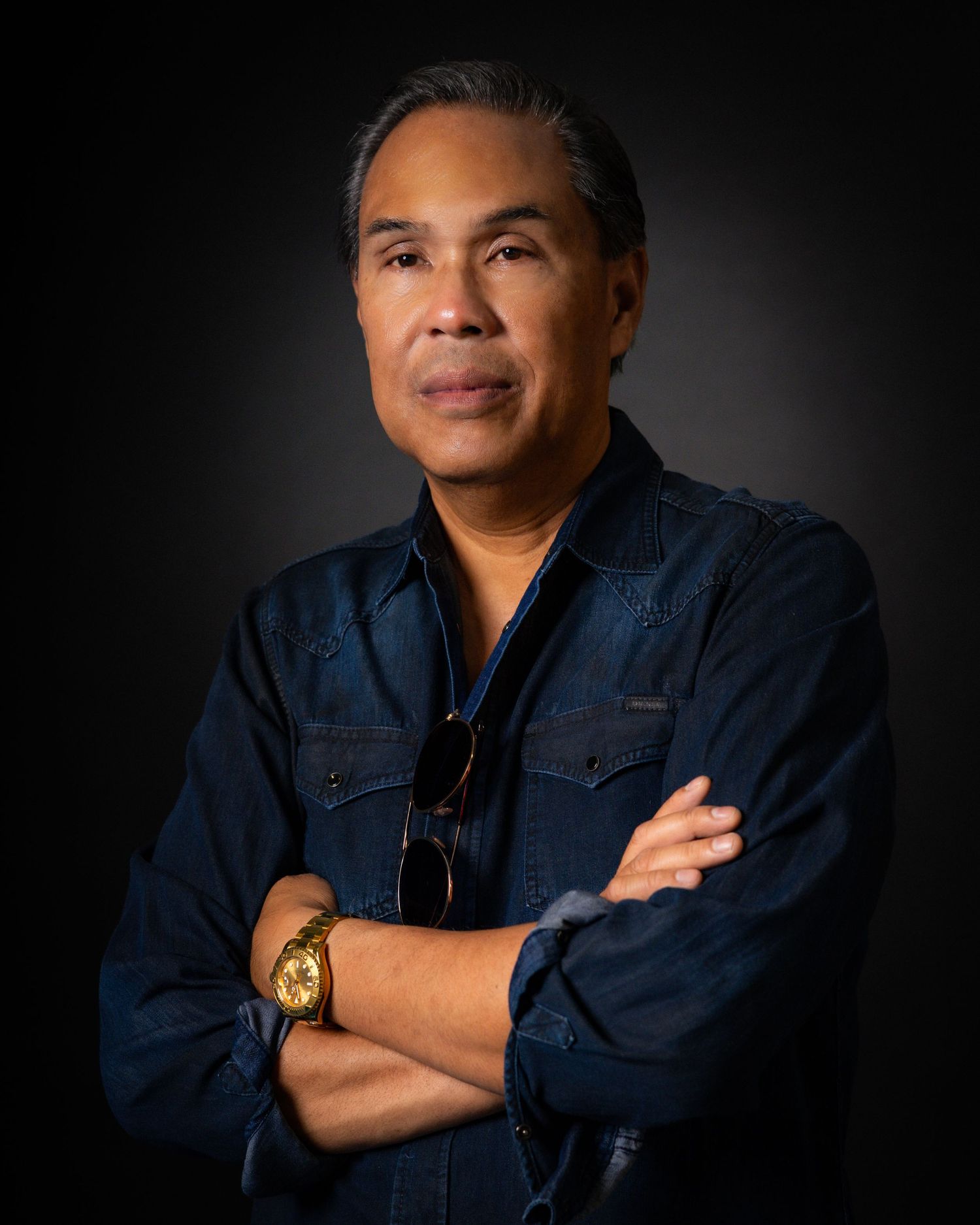 A confident man in a blue denim shirt with arms crossed, wearing a gold watch and sunglasses tucked in collar.