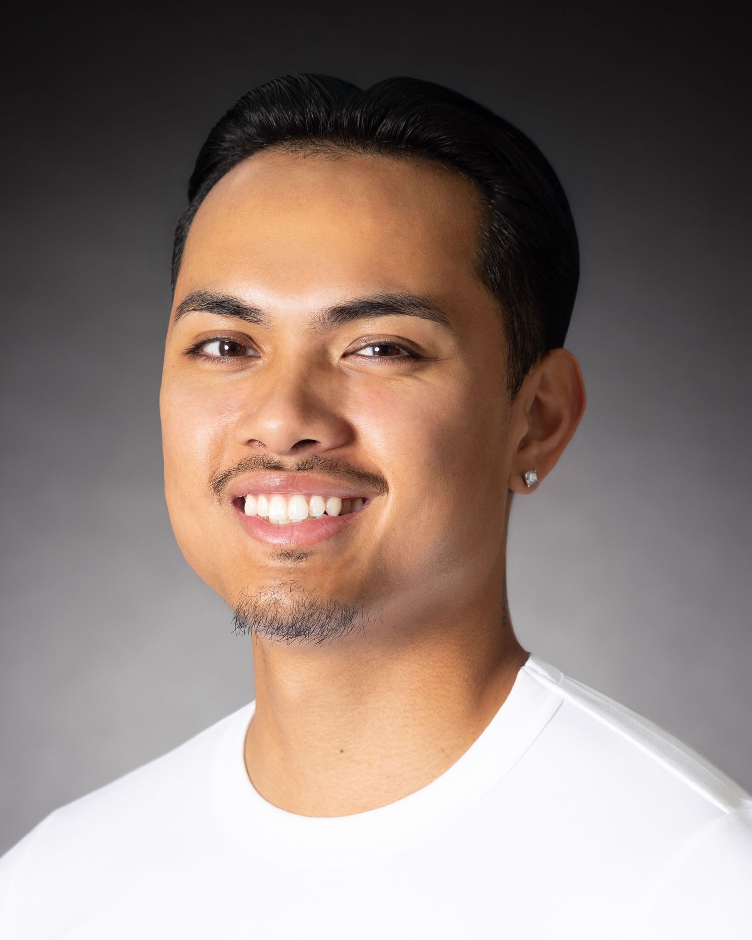 Young man with short dark hair, goatee, and ear stud smiles confidently in a white shirt against gray background.