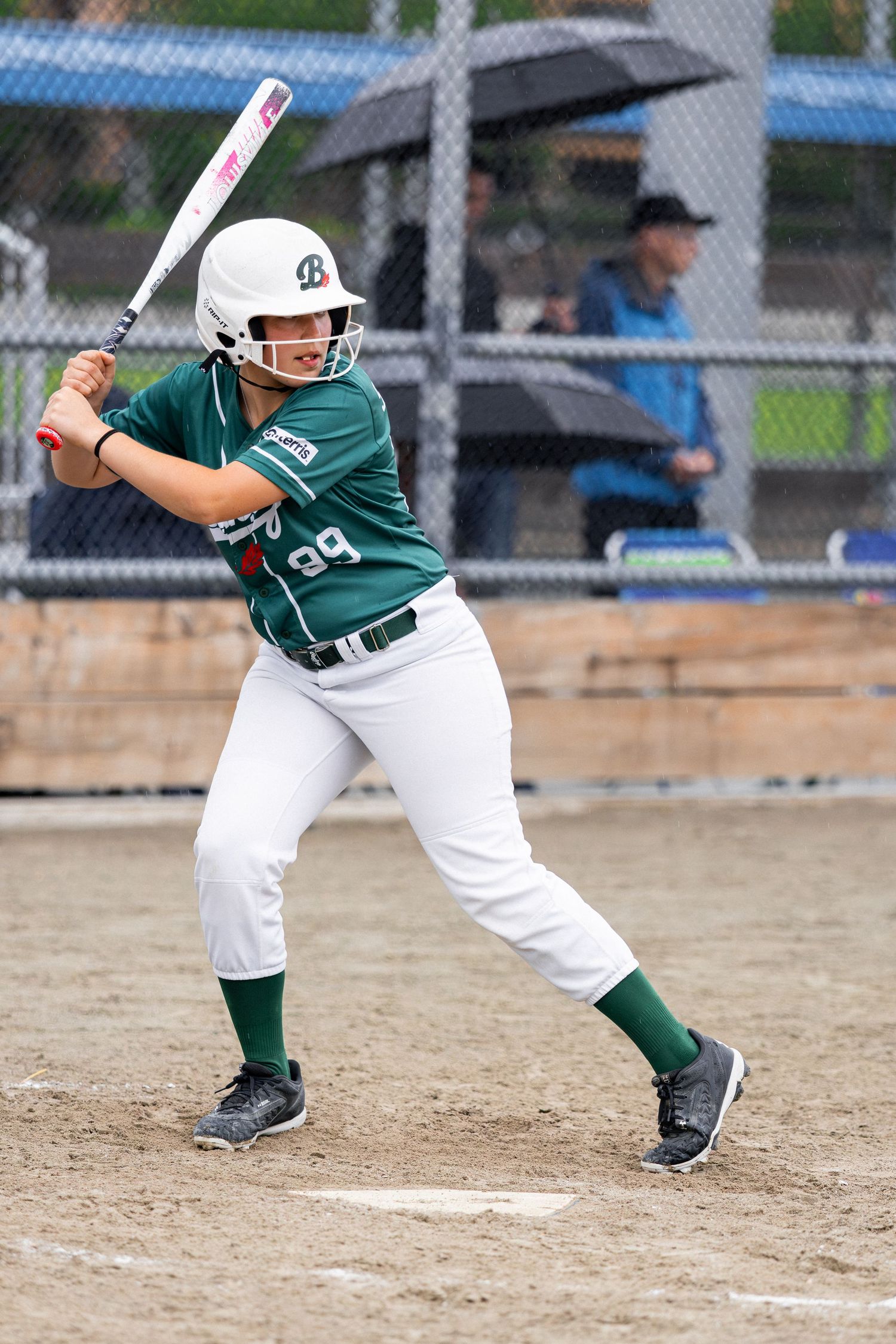 Young softball player in green uniform #99 stands ready at bat during a rainy game.