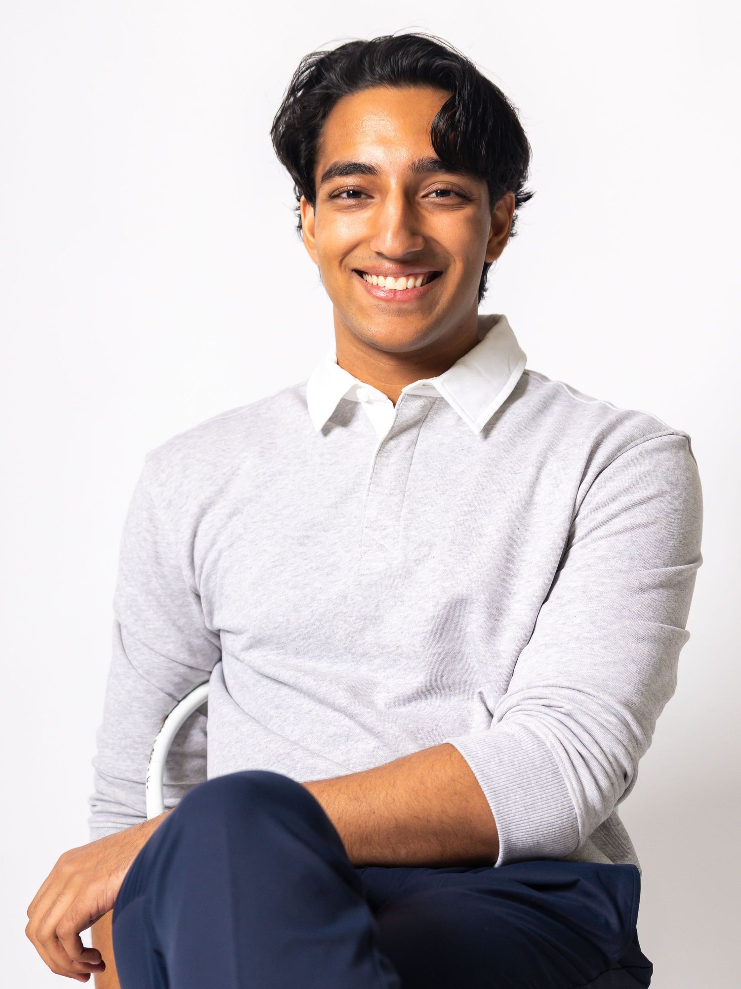 Smiling young man in a gray long-sleeve polo shirt and navy pants, seated casually against a white background.