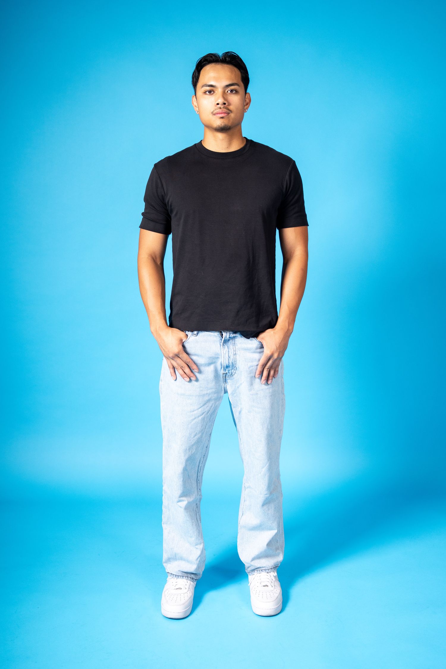 Young man in black t-shirt and light wash jeans posing against a bright blue background with hands in pockets.