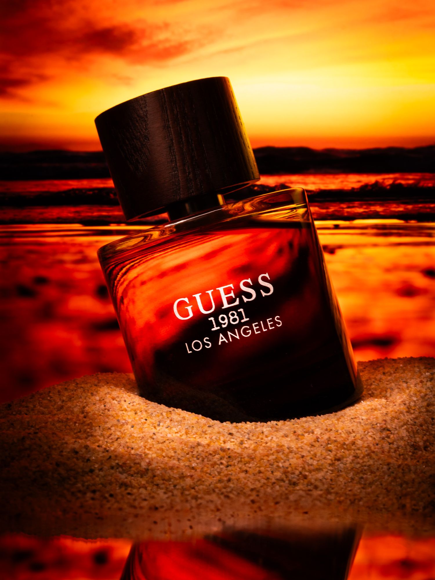Guess 1981 Los Angeles cologne bottle resting in sand against a dramatic orange sunset sky.