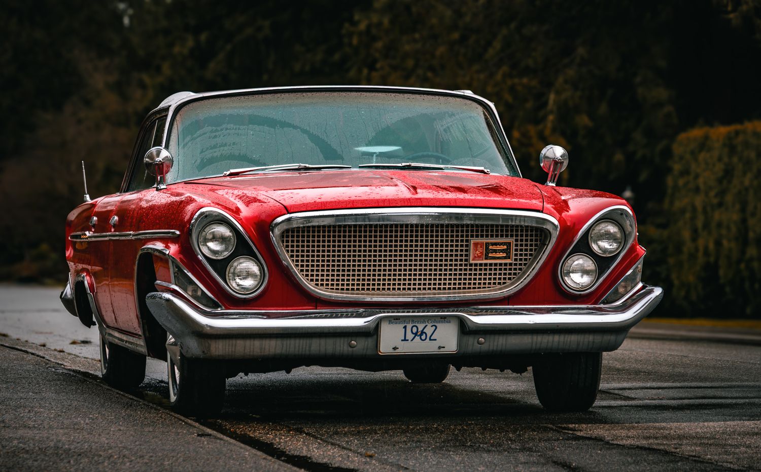 A classic red 1962 Chrysler vintage car parked on a road surrounded by lush green trees.
