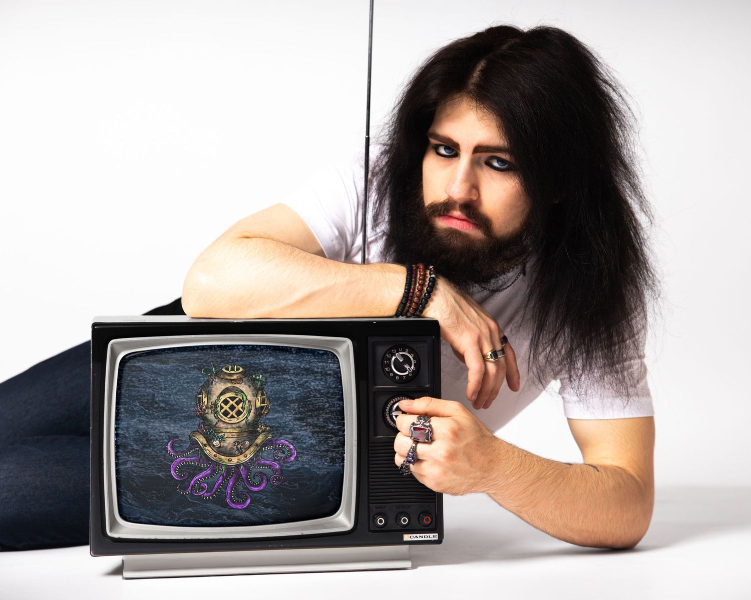 Bearded man with long dark hair poses beside a vintage TV displaying an octopus emblem on a white background.