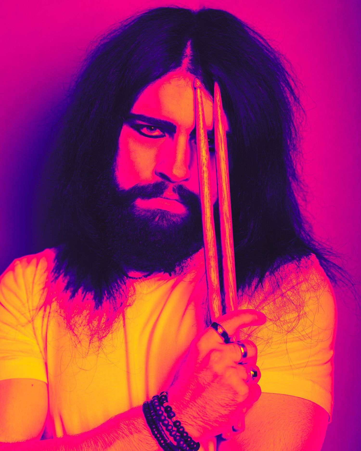 Bearded musician with long dark hair holds drumsticks, bathed in vibrant pink and yellow neon lighting.