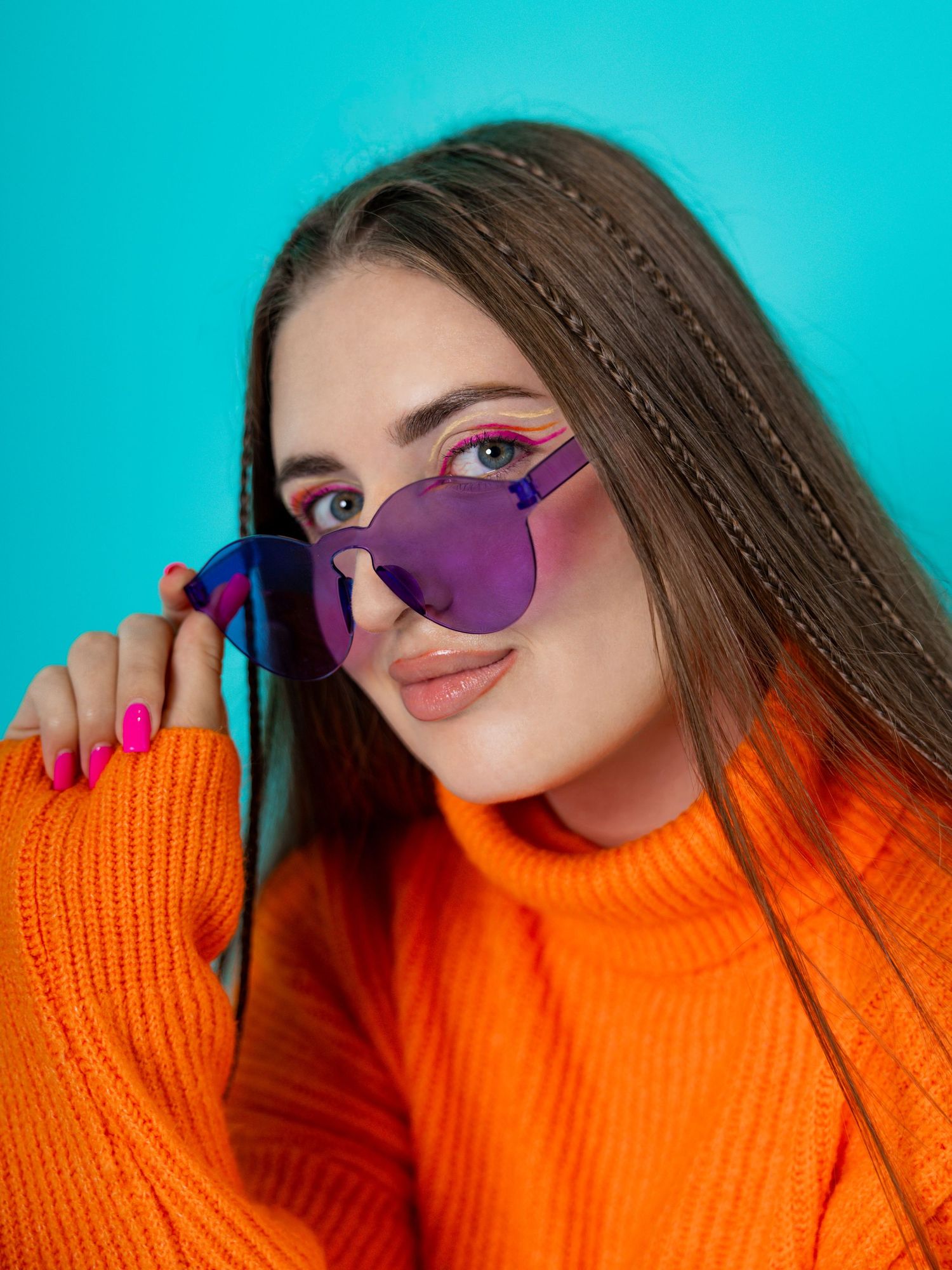 Young woman in orange sweater wearing purple heart-shaped sunglasses against a teal background.