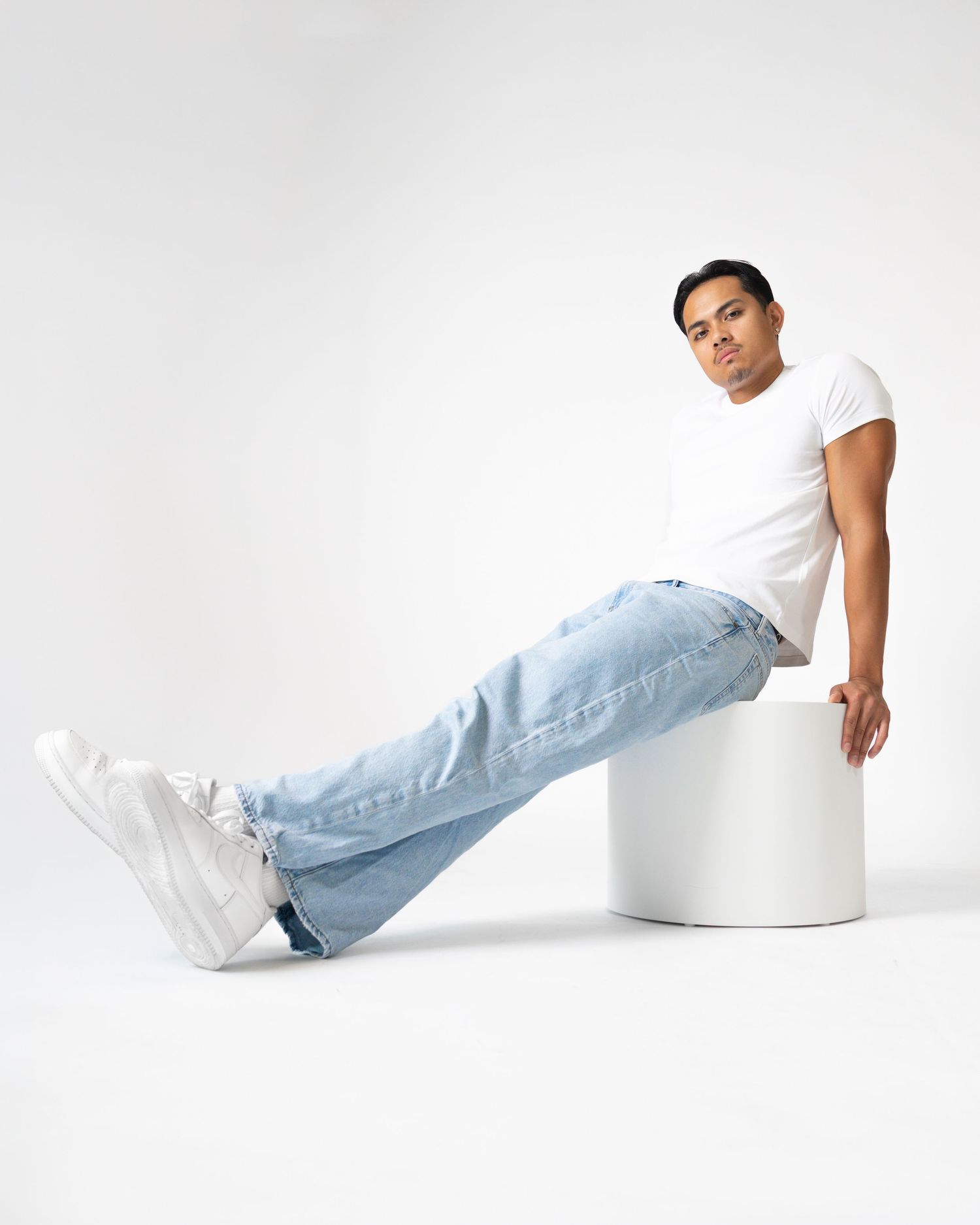 Man in white t-shirt and light blue jeans sitting on white cylinder pedestal against white background.