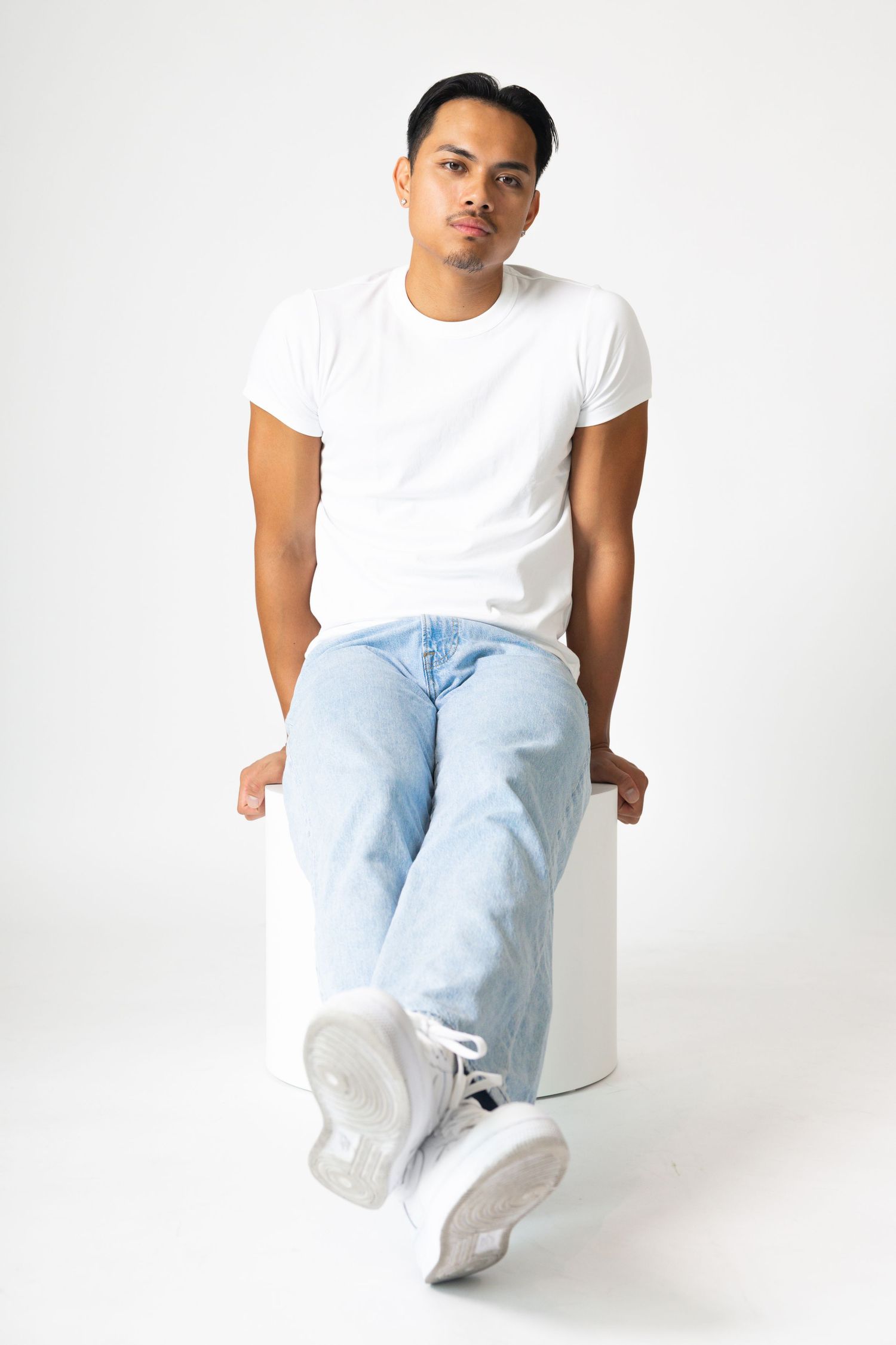 Young man sitting on white cube wearing white t-shirt, light blue jeans, and white sneakers against white background.